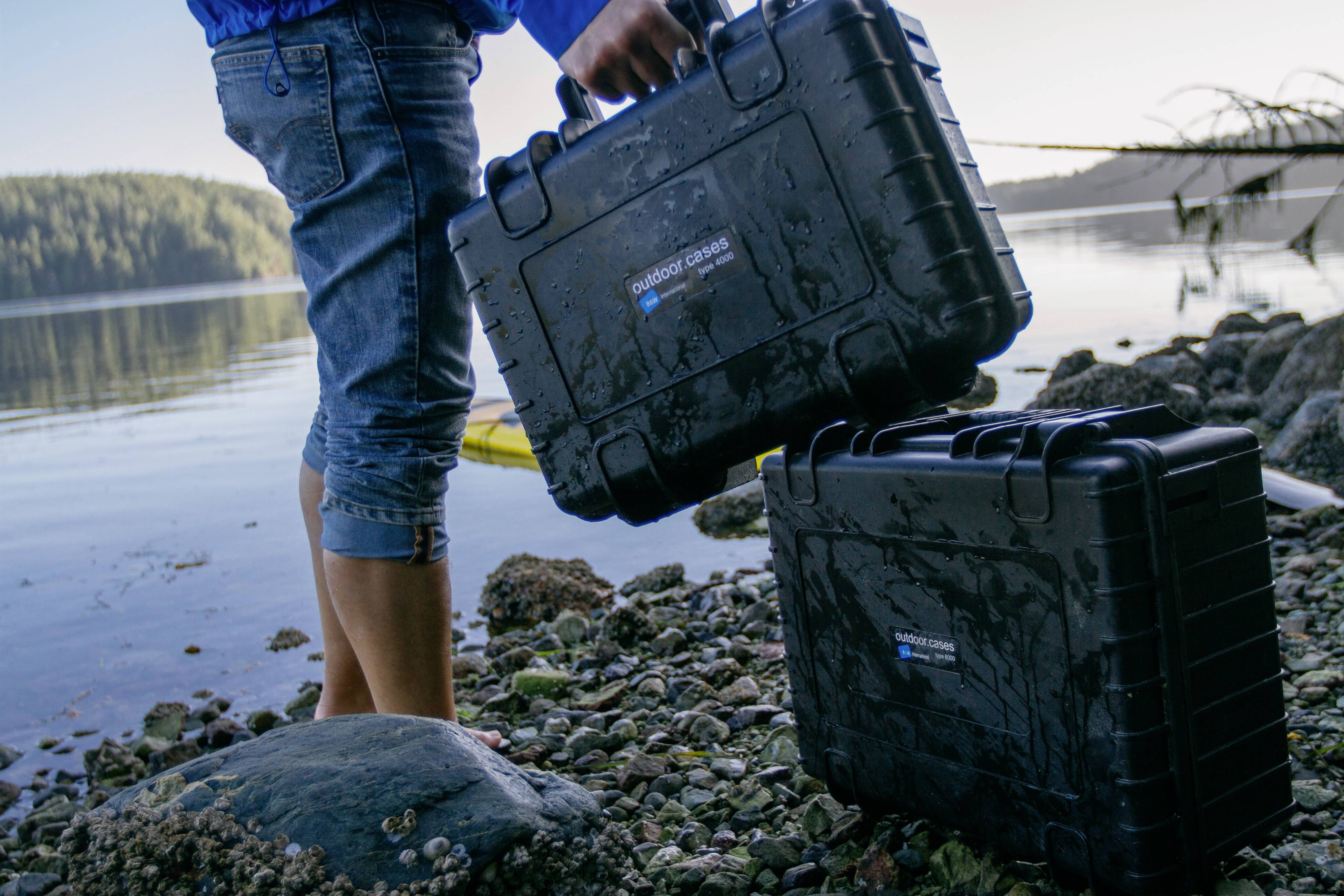 Eine Person am Wasser trägt einen wasserdichten schwarzen Koffer. Ein zweiter Koffer liegt auf dem Kies. Hintergrund: See und Bäume.