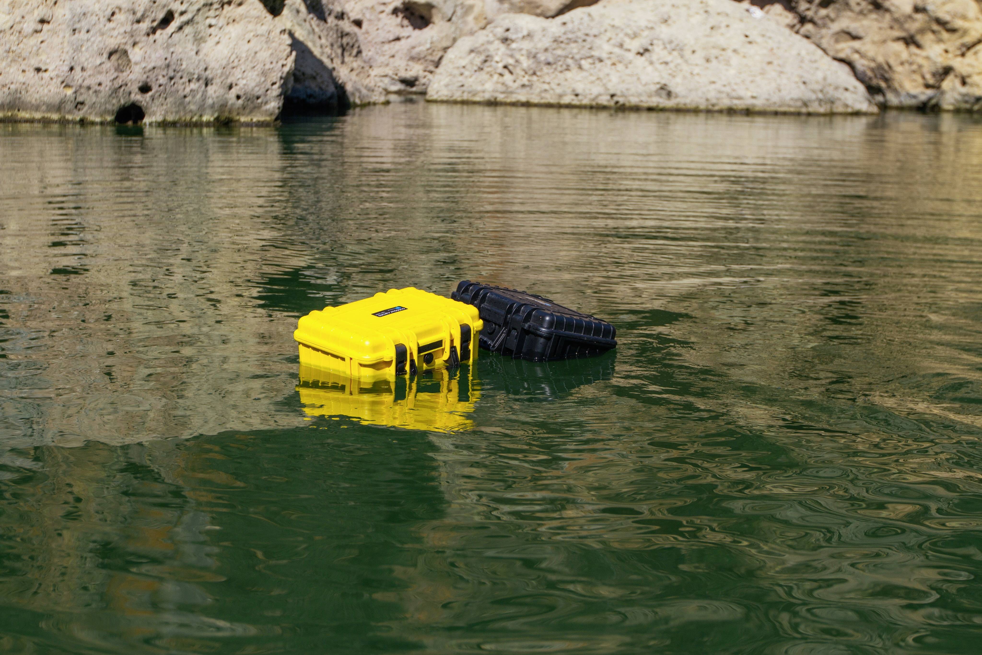 Zwei schwimmende Sicherheitskoffer, ein gelber und ein schwarzer, treiben auf einem grünen Gewässer, umgeben von Felsen.
