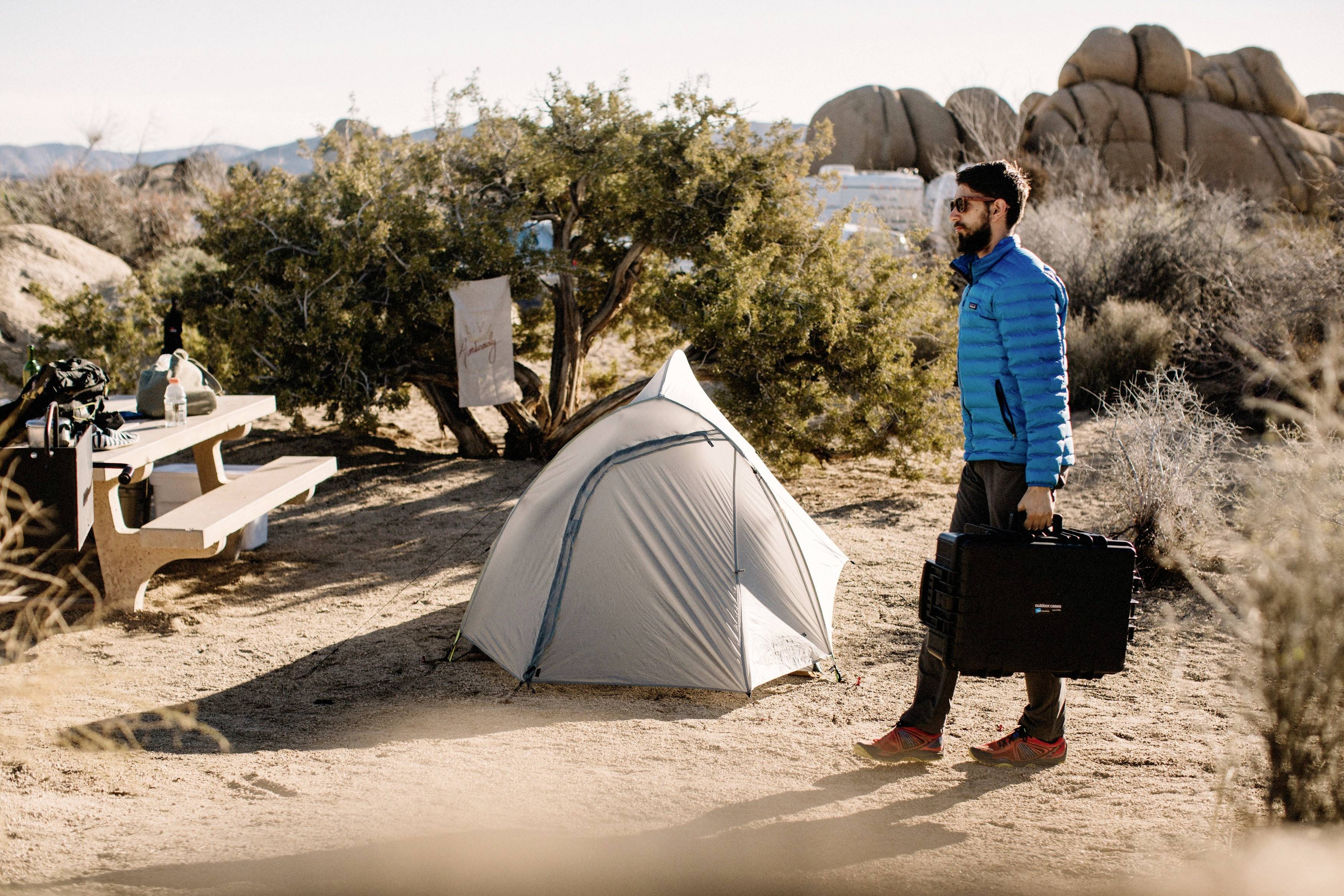 Ein Mann in blauer Jacke trägt einen Koffer an einem Zeltplatz mit Zelten und Bäumen; sonniger Tag mit Felsen im Hintergrund.