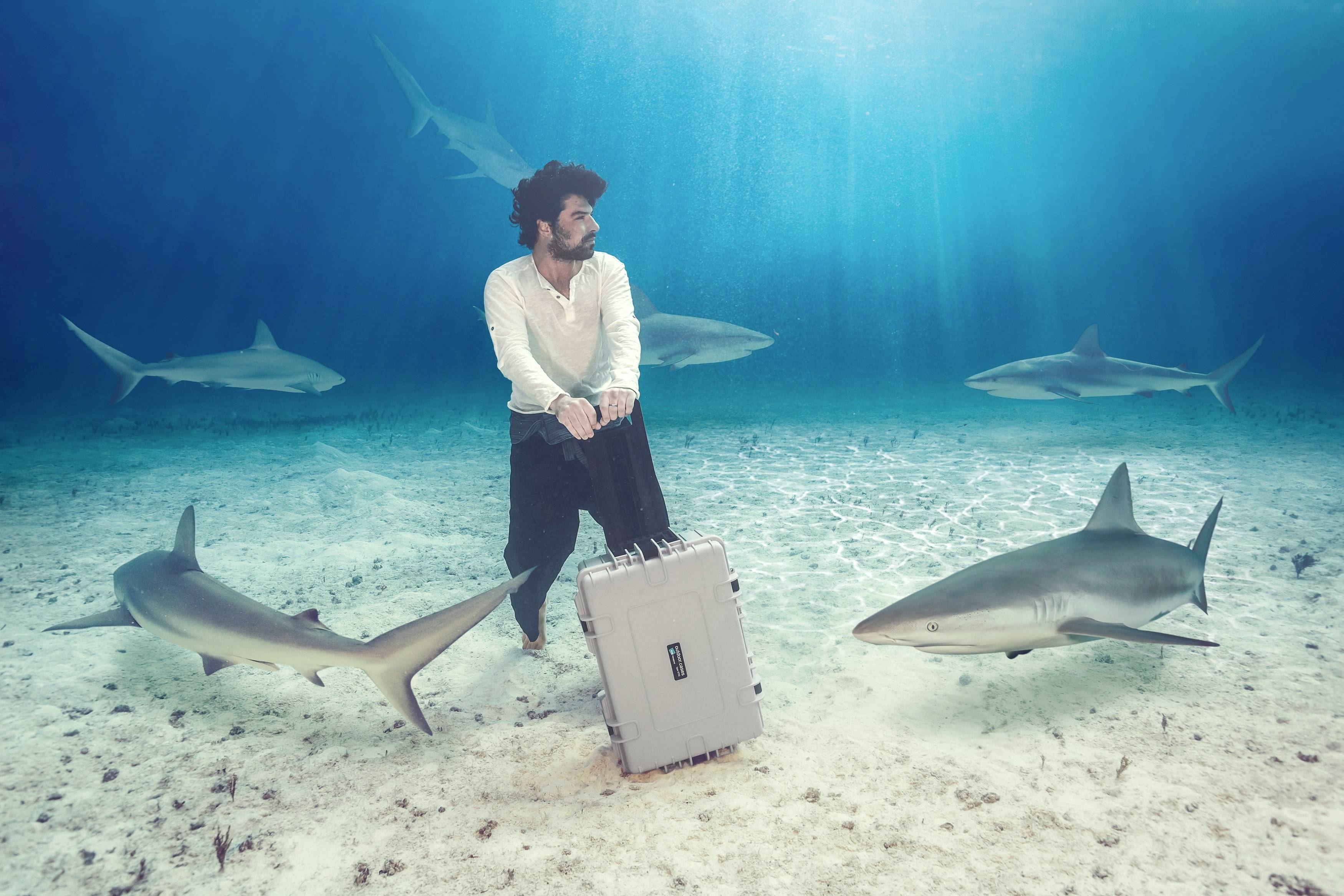 Ein Mann steht unter Wasser neben einem Koffer, umgeben von Haien. Die Szene wirkt surreal und inszeniert.