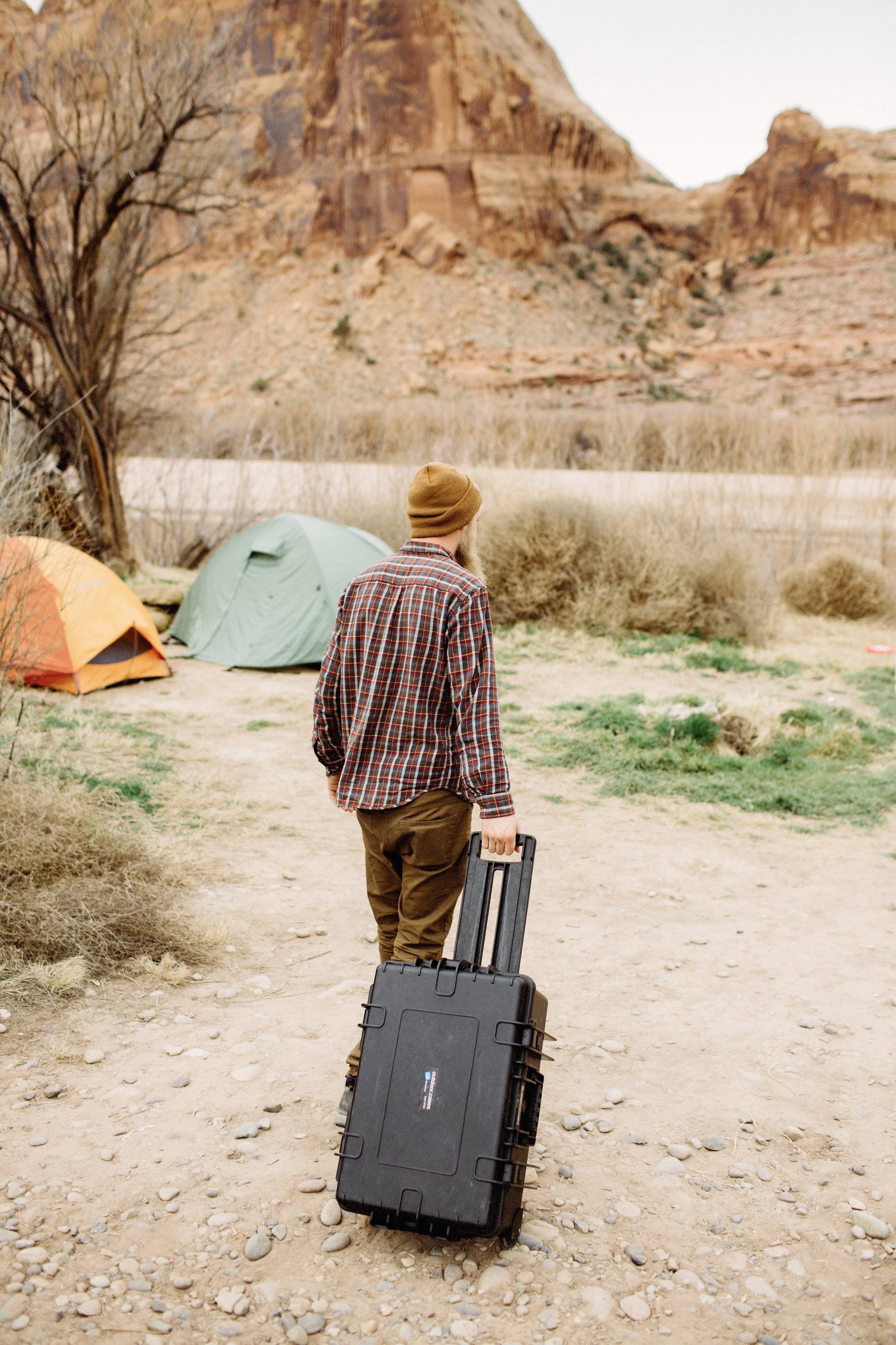 Person mit braunem Hut zieht einen schwarzen Rollkoffer in einem Campinggebiet mit Zelten und felsiger Landschaft im Hintergrund.
