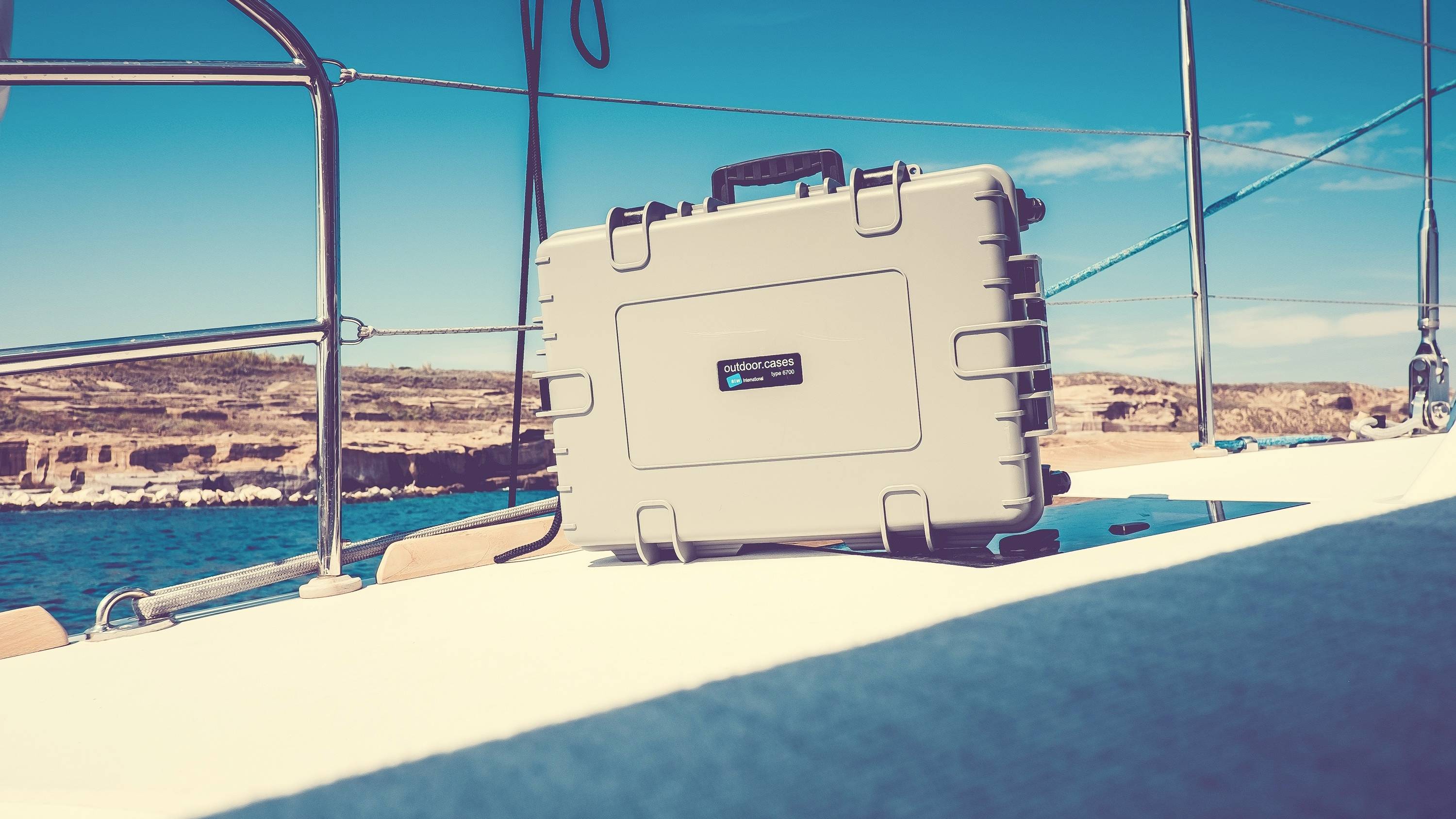 Ein silberner Koffer liegt auf dem Deck eines Segelboots, das auf einem ruhigen Meer segelt. Im Hintergrund ist die Küste zu sehen.
