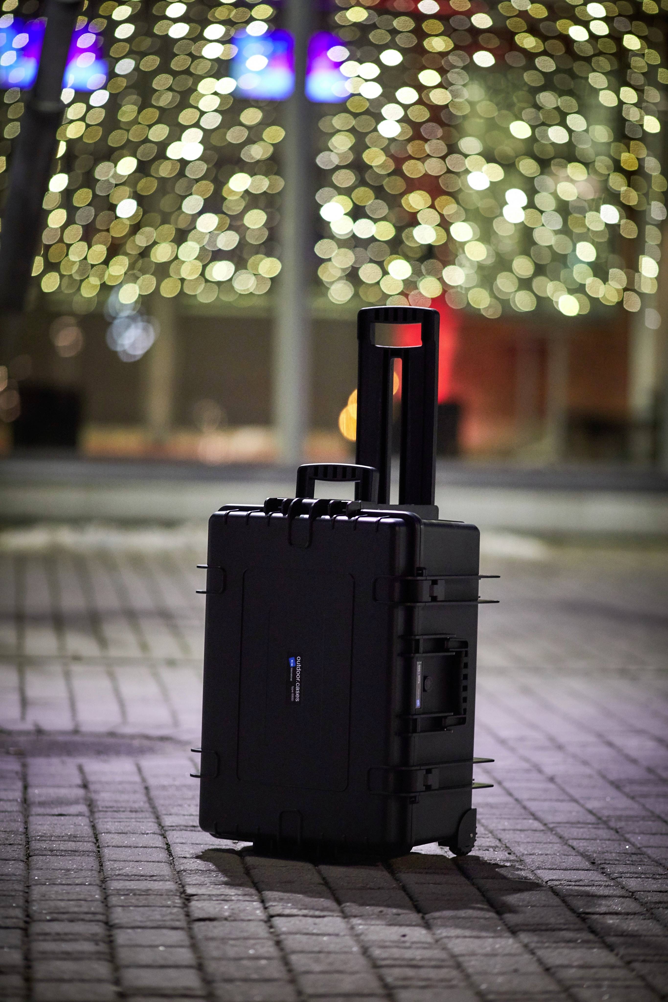 Ein schwarzer Rollkoffer steht auf einem gepflasterten Weg. Im Hintergrund sind unscharfe Lichter zu sehen, die eine stimmungsvolle Beleuchtung erzeugen.