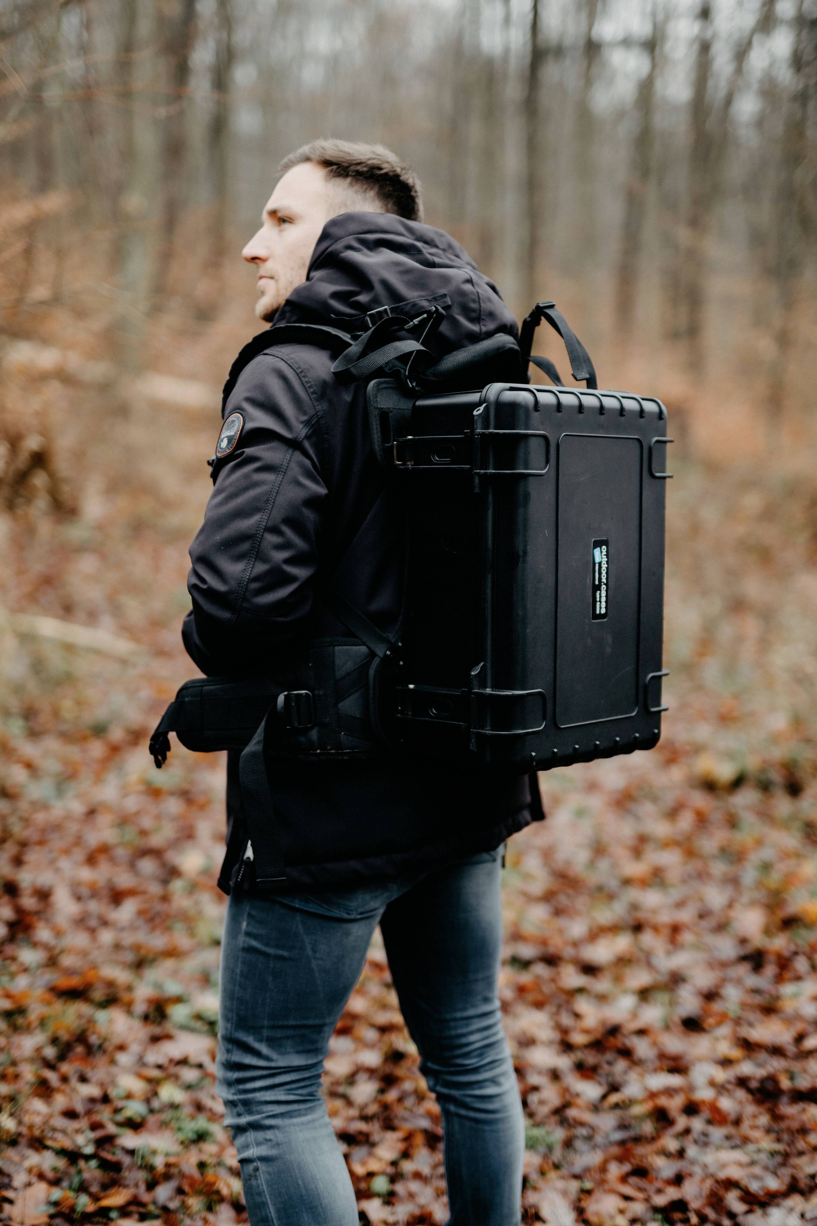 Ein Mann im Wald trägt einen großen schwarzen Rucksackkoffer. Es ist Herbst, die Blätter sind braun und der Himmel bewölkt.