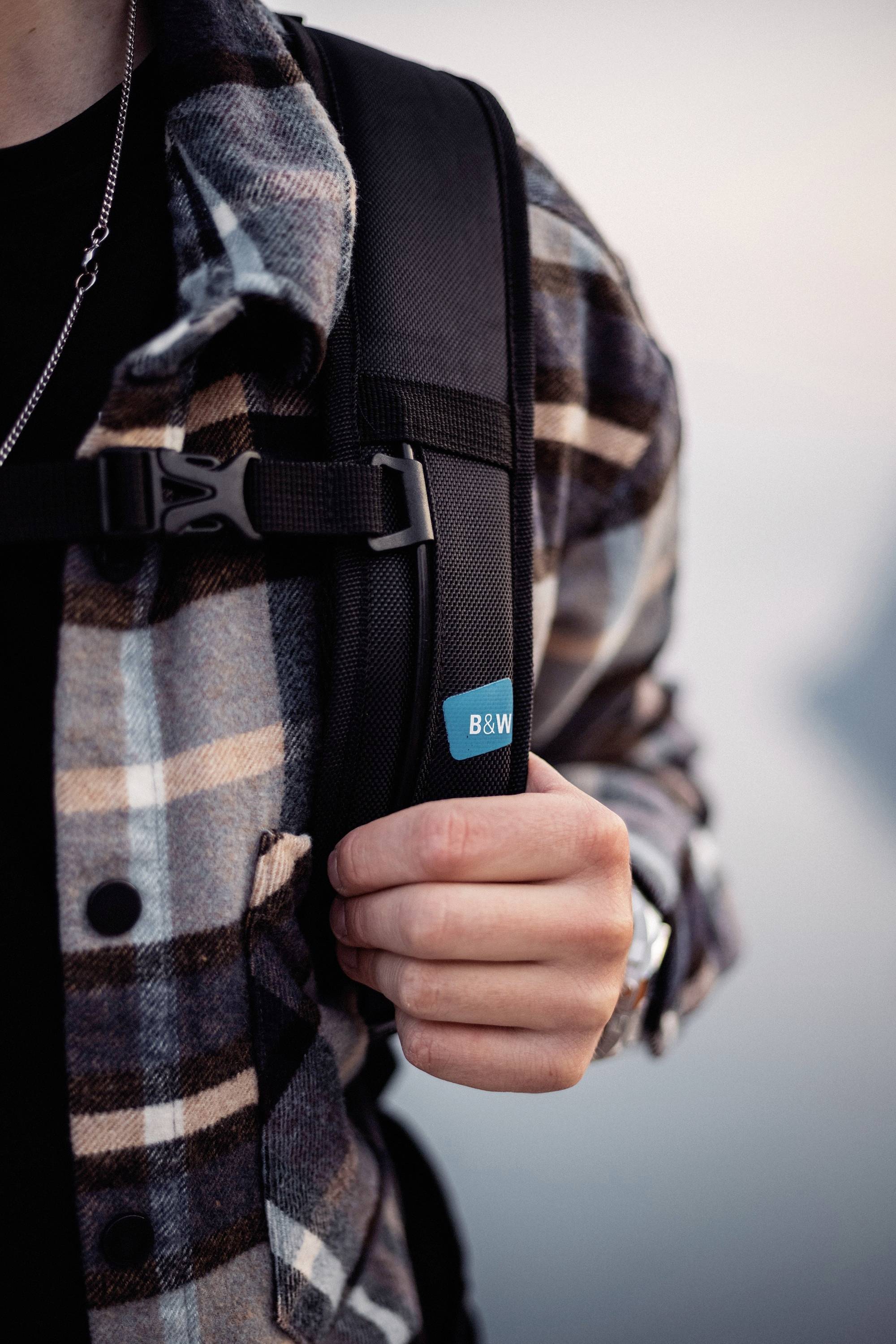 Person trägt kariertes Hemd und schwarzen Rucksack, hält Träger mit rechter Hand. Atmosphärische Szene, unscharfer Hintergrund.