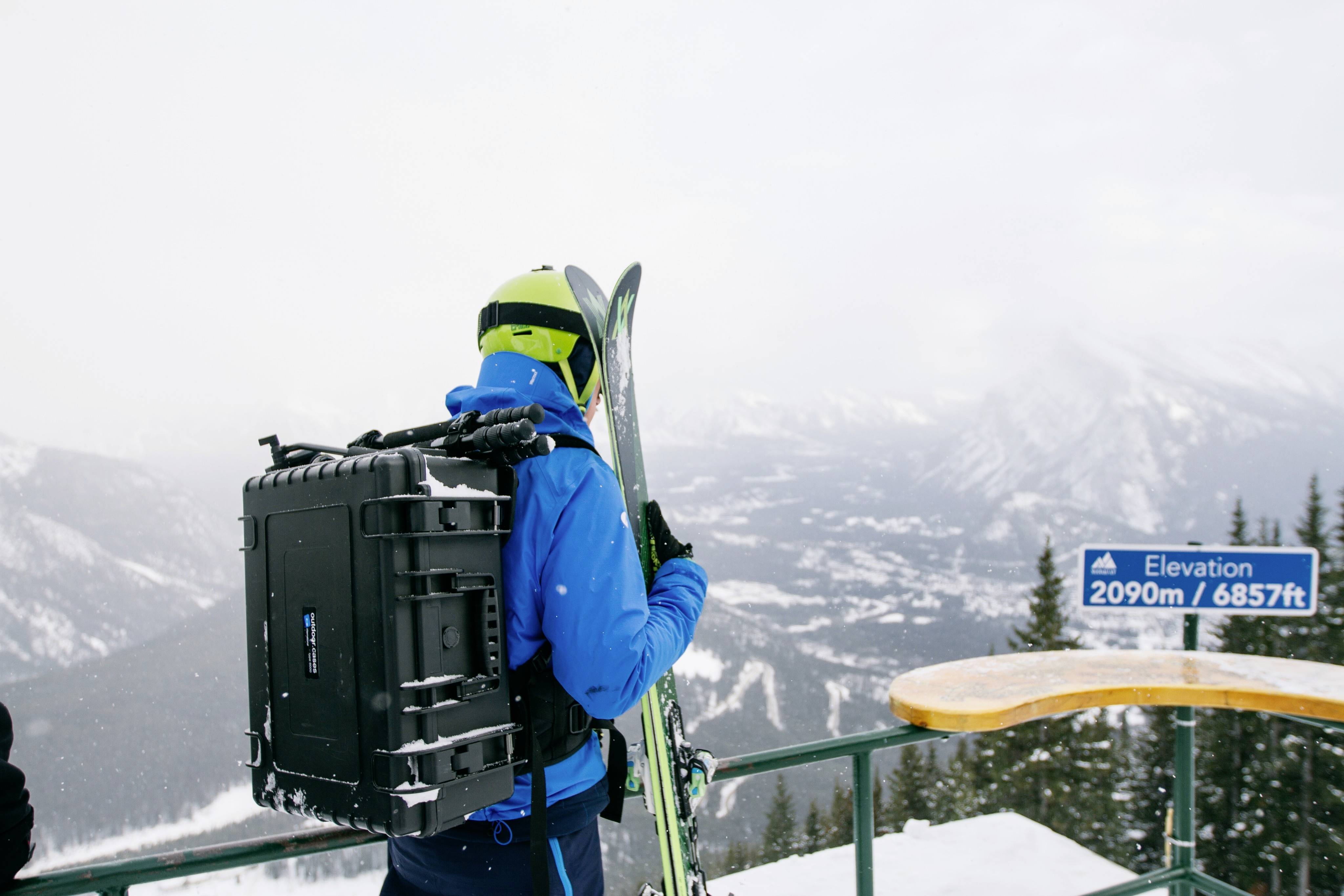 Eine Person in blauer Skiausrüstung steht auf einem verschneiten Berg mit Skiern und Ausrüstung, Blick auf bergige Landschaft und eine Höhenangabe: '2090m / 6857ft'.