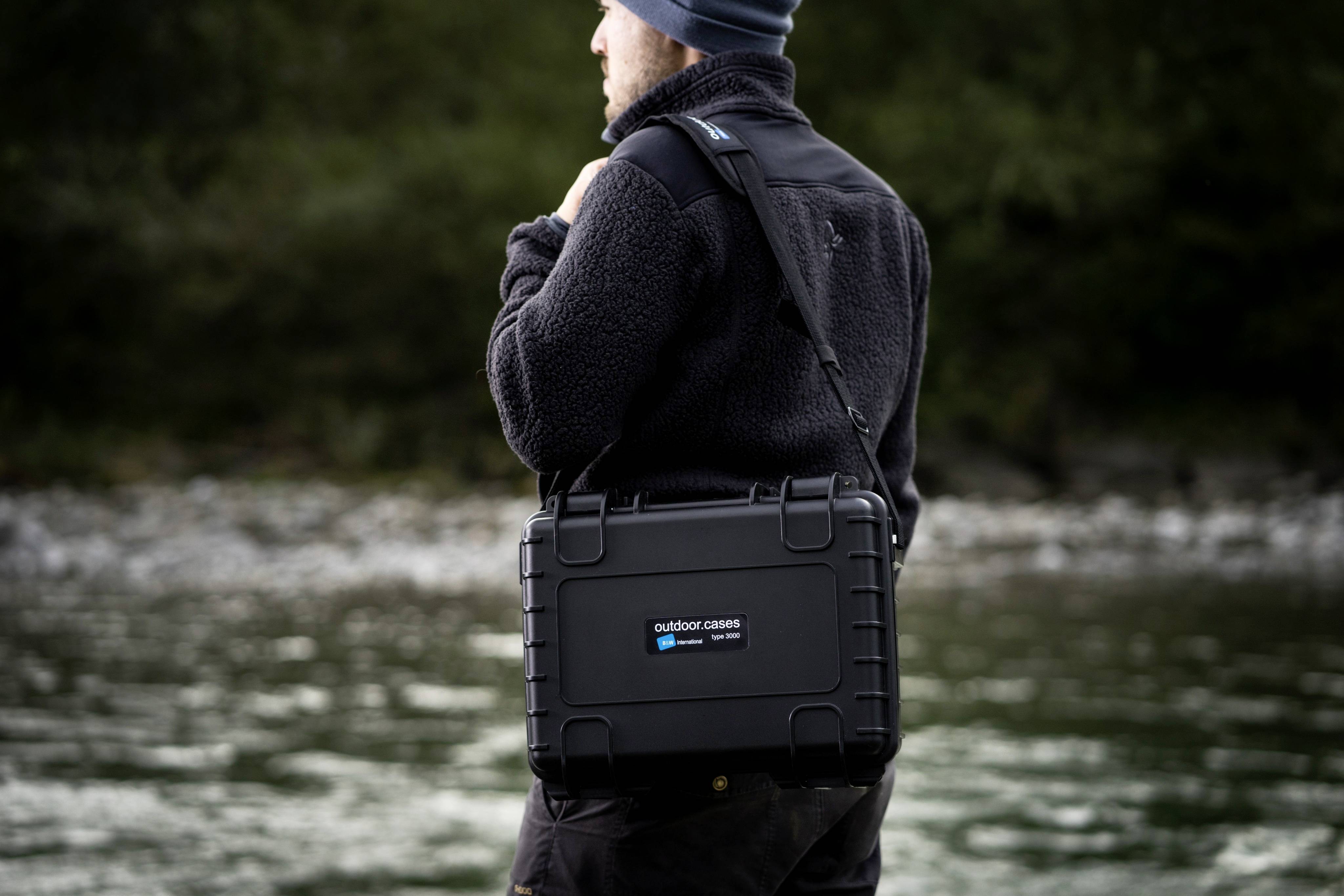 Eine Person im schwarzen Outdoorkleidung trägt eine robuste, schwarze Schutzkiste mit der Aufschrift 'outdoor cases' am Ufer eines Flusses.