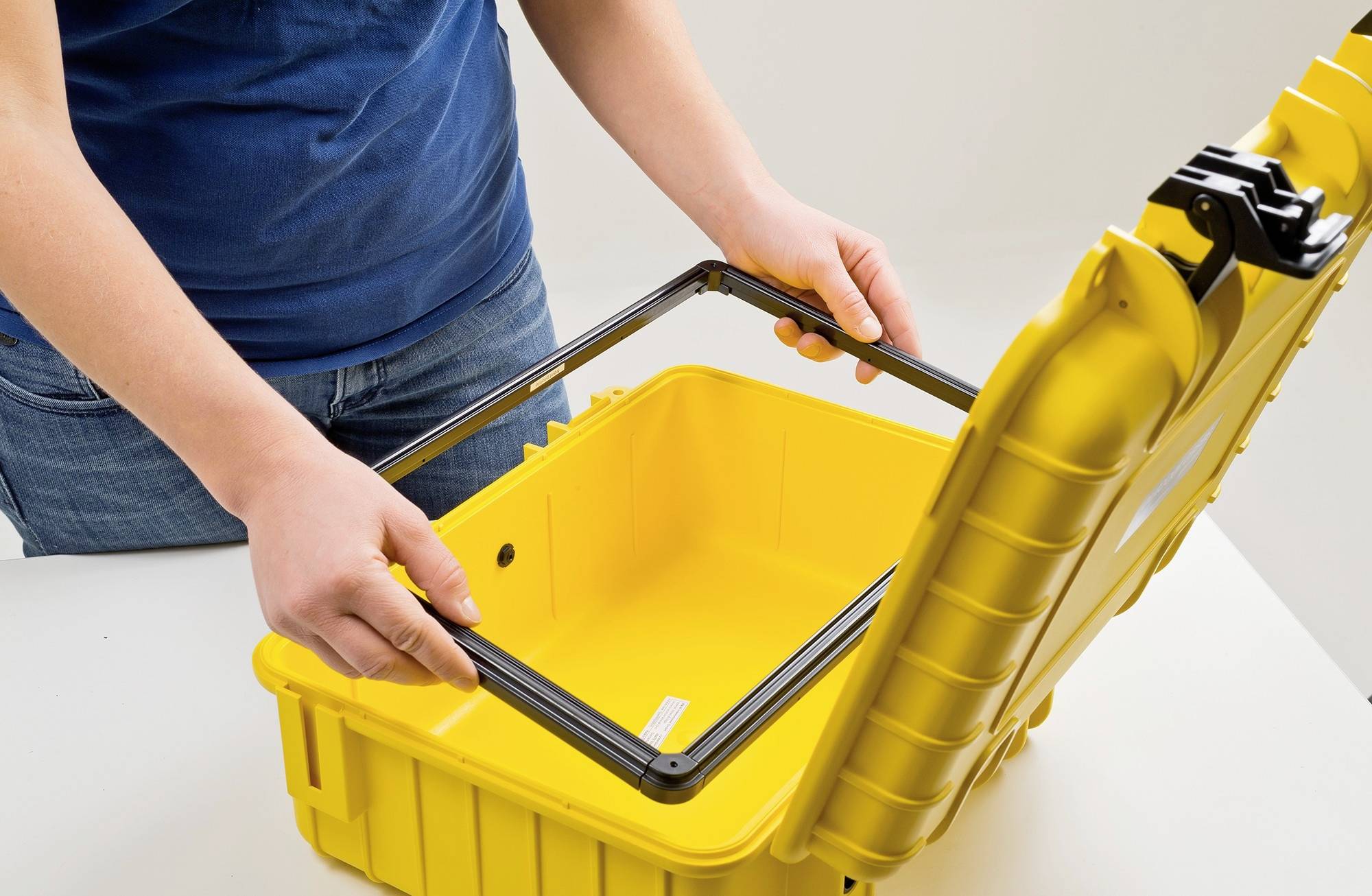 Eine Person öffnet einen gelben Schutzkoffer mit einem schwarzen Rahmen. Der Koffer ist leer und steht auf einem Tisch.