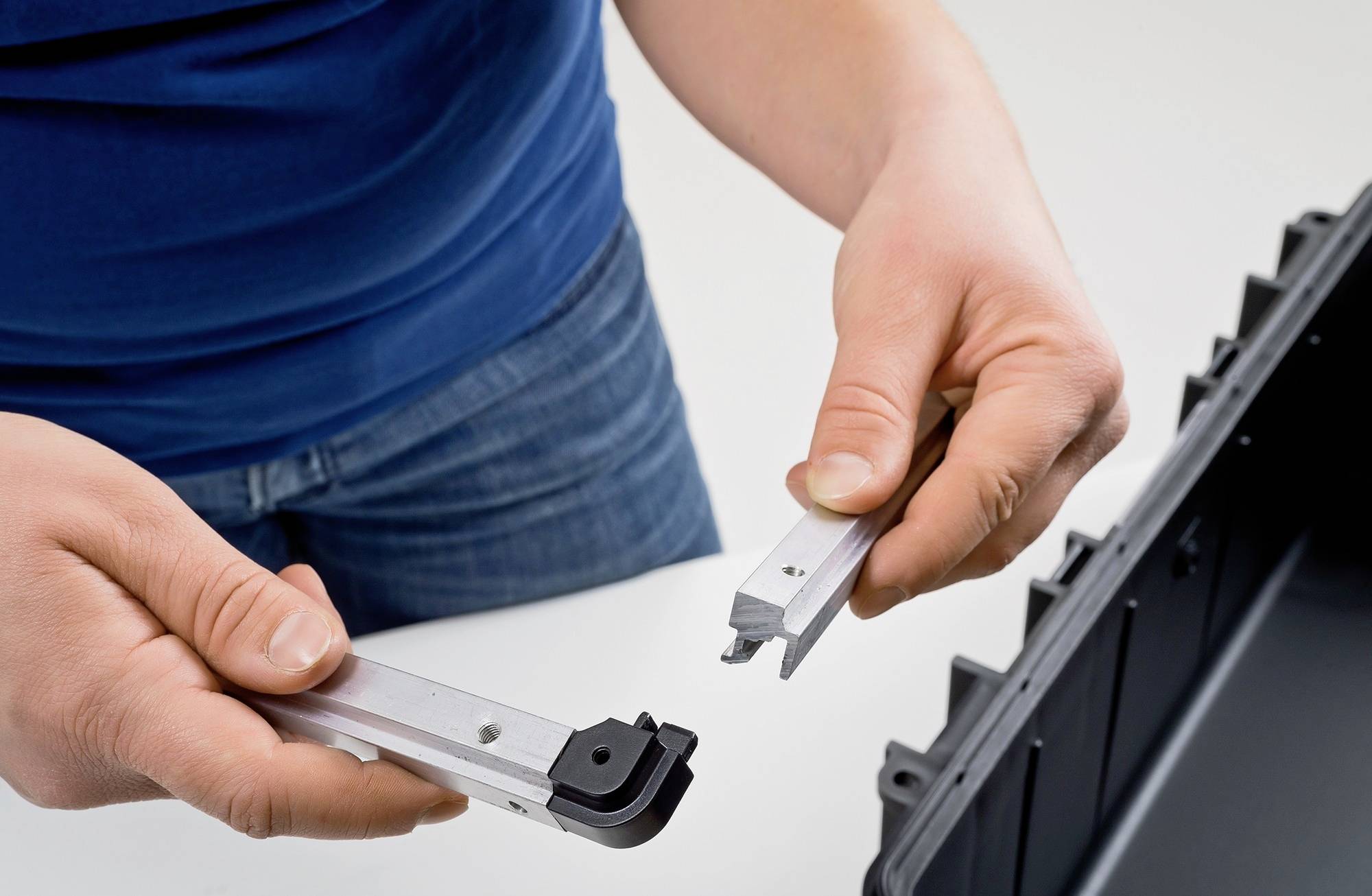 Eine Person montiert ein Metallteil an einem schwarzen Kunststoffbehälter. Hände halten zwei silberne Metallstücke.