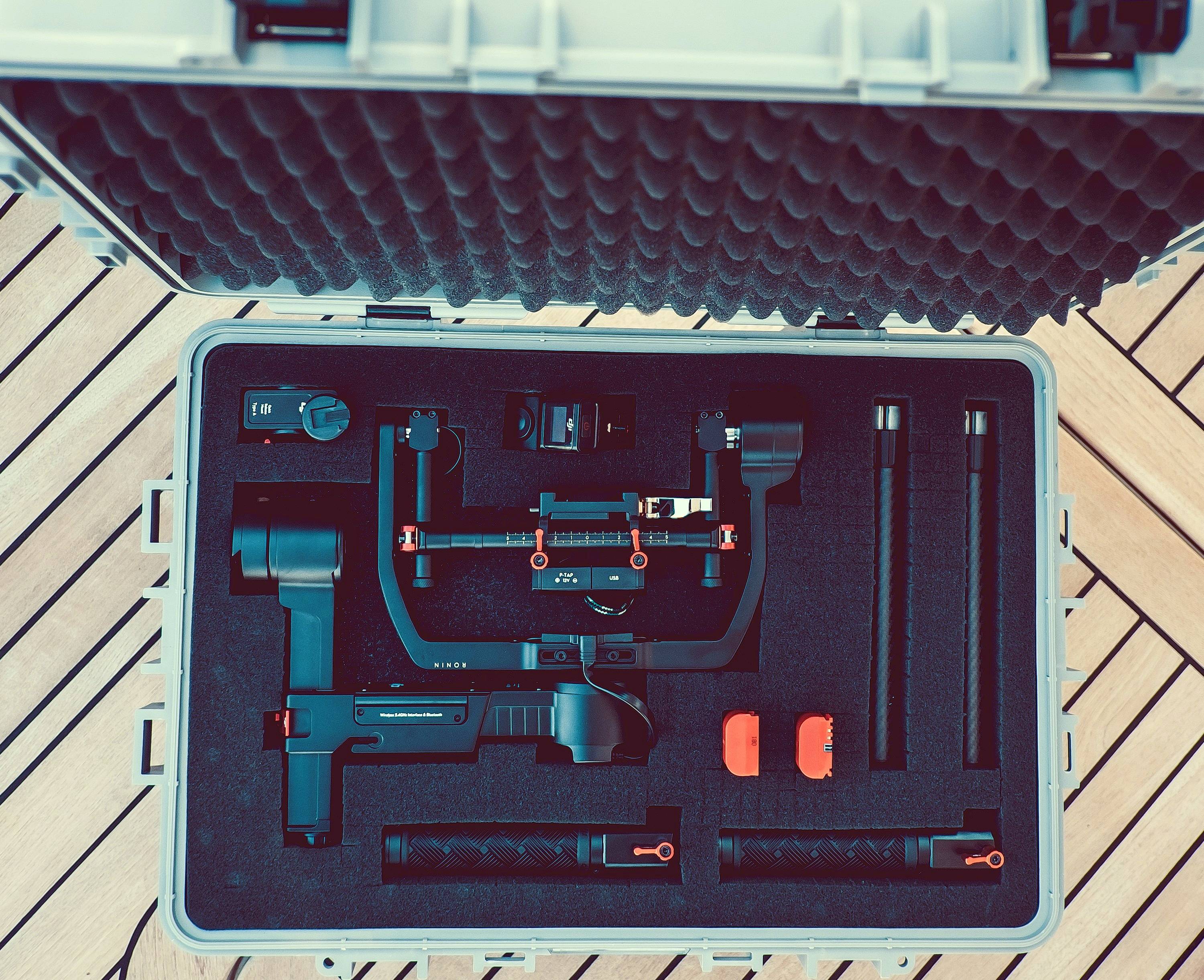 Ein geöffneter Koffer zeigt ein elektronisches Stabilisierungssystem mit Griffen und Zubehör. Der Koffer liegt auf einem Holzdeck.