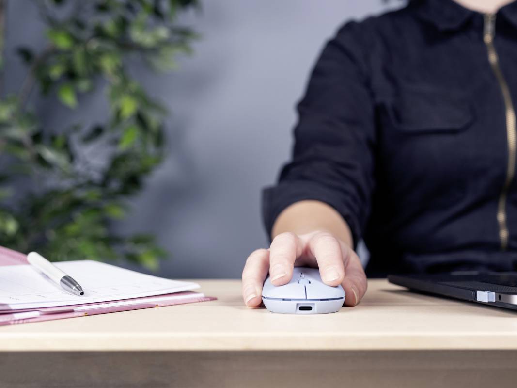 Eine Person in schwarzer Kleidung benutzt eine kabellose Maus auf einem Tisch. Daneben liegen ein Notizbuch mit Stift und ein Laptop.