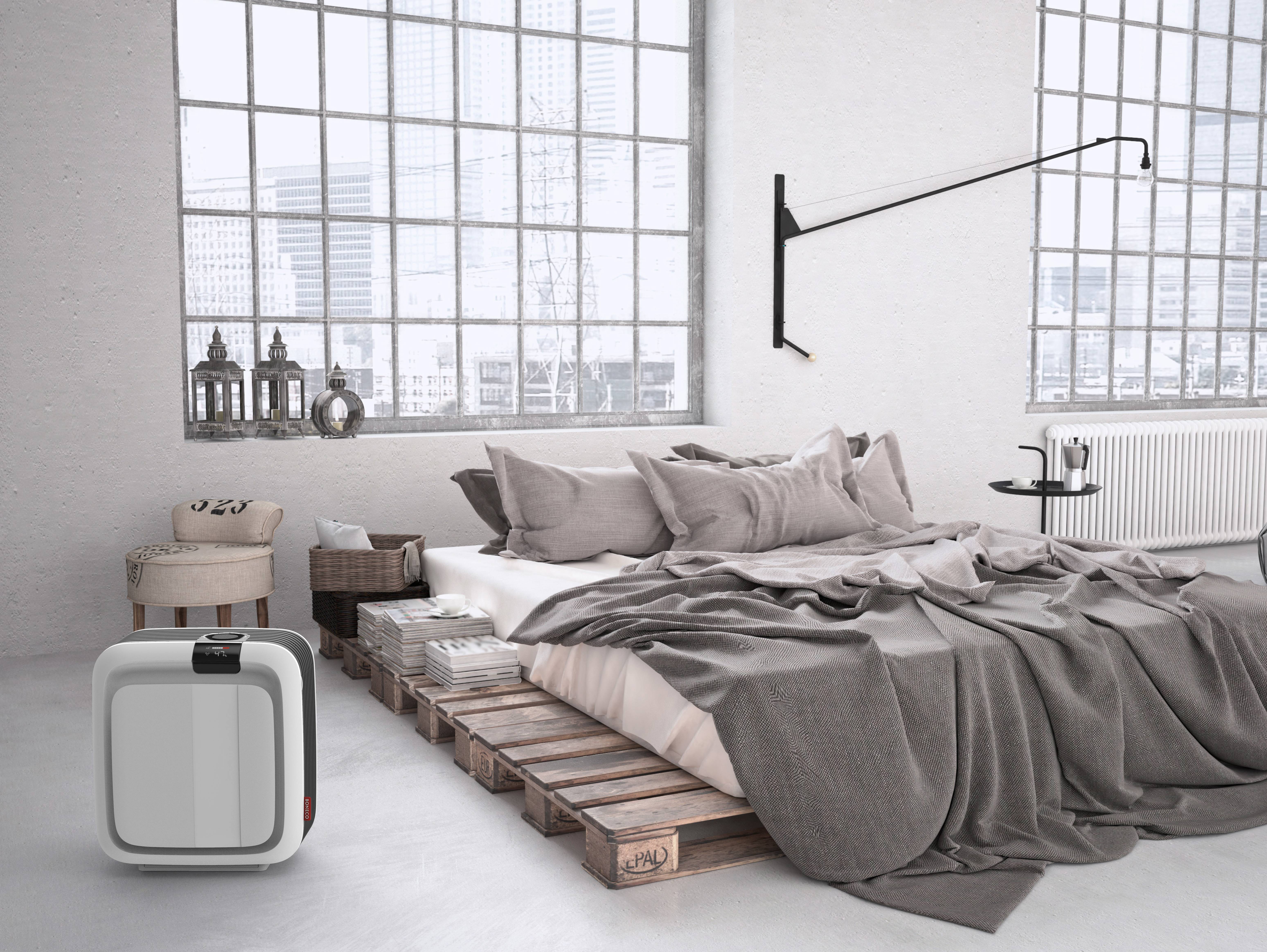 Ein modernes Schlafzimmer mit einem Bett aus Paletten, grauer Bettwäsche und großen Fenstern. Ein dekorativer Heizkörper und eine minimalistische Wandlampe ergänzen das Design.