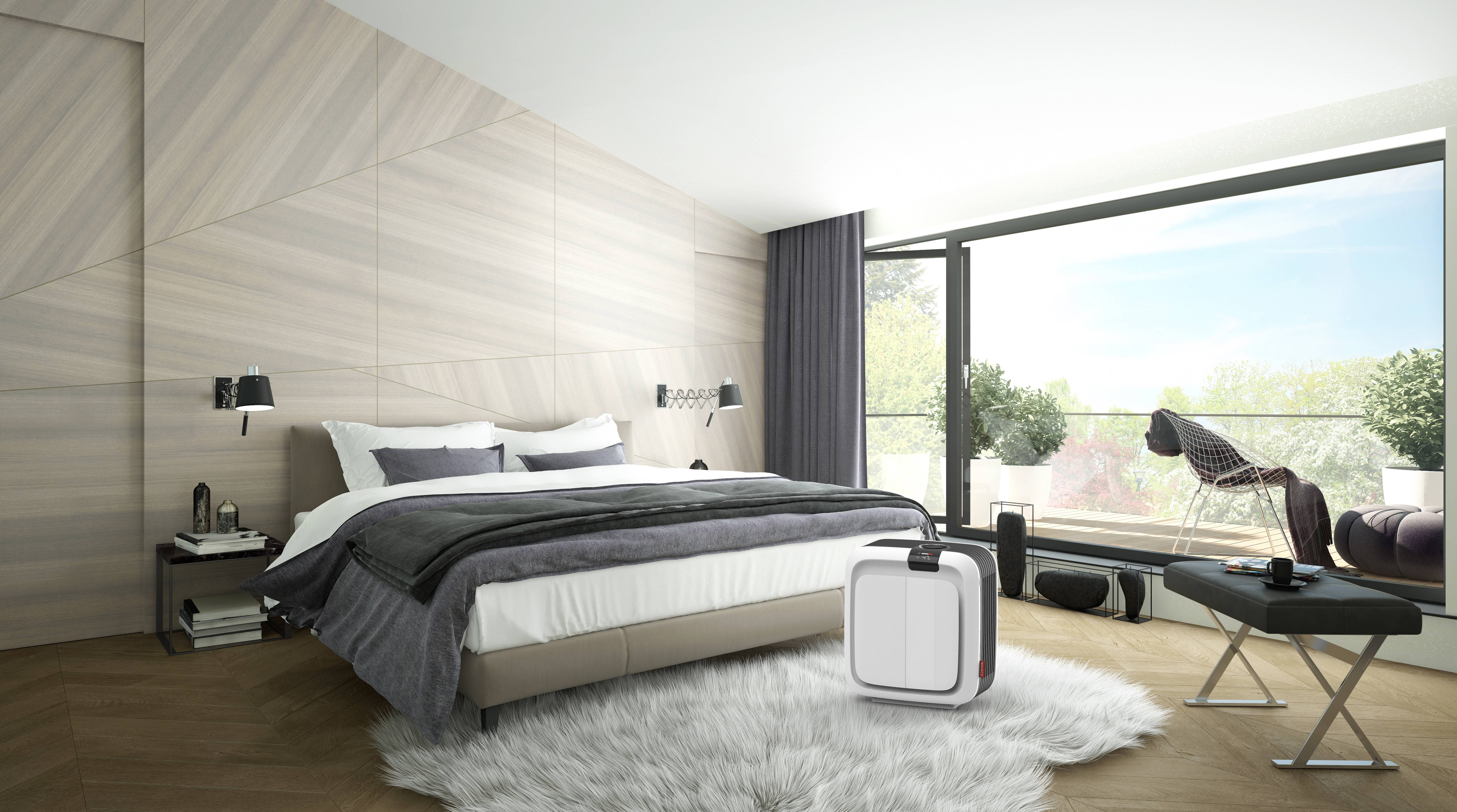 Ein modernes Schlafzimmer mit einem großen Bett, grauen Decken, einem Luftreiniger auf dem Boden und Blick auf einen sonnigen Balkon.