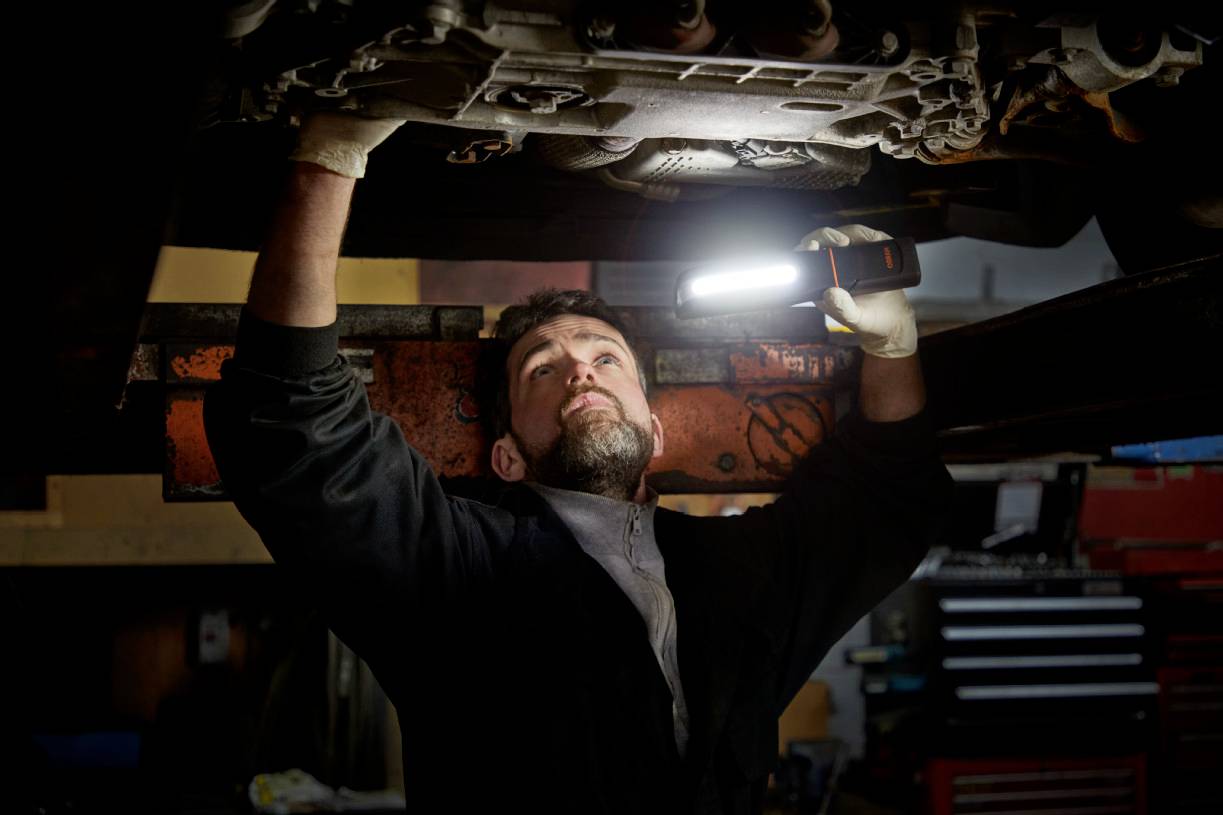 Ein Mechaniker repariert unter einem Fahrzeug und beleuchtet den Bereich mit einer tragbaren Lampe, um besser sehen zu können.