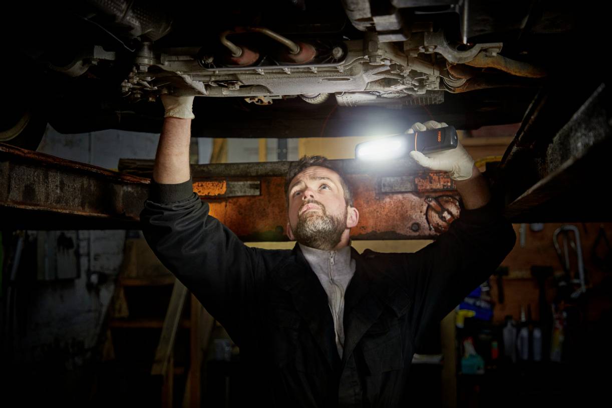 Ein Mechaniker arbeitet unter einem Auto und leuchtet mit einer Taschenlampe in den Motorraum einer Werkstattumgebung.