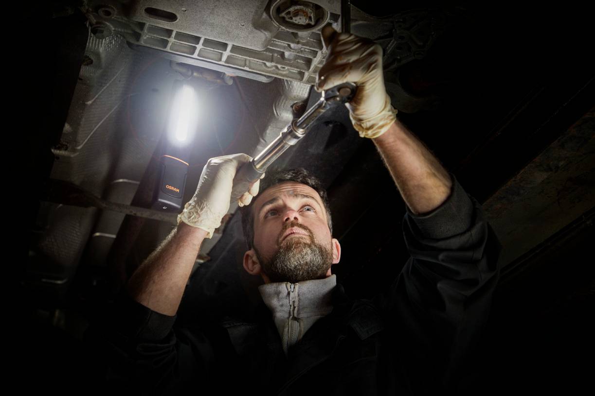 Mechaniker arbeitet unter einem Auto, hält einen Schraubenschlüssel und trägt Handschuhe. Eine Lampe beleuchtet den Arbeitsbereich.