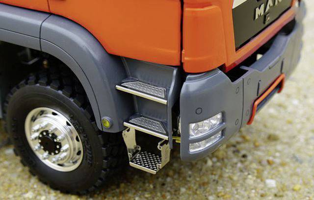 Nahaufnahme eines orangefarbenen Lastwagens mit gut sichtbaren Trittstufen und Reifen auf einer Kiesfläche. Der Fokus liegt auf den Details der Trittstufen.