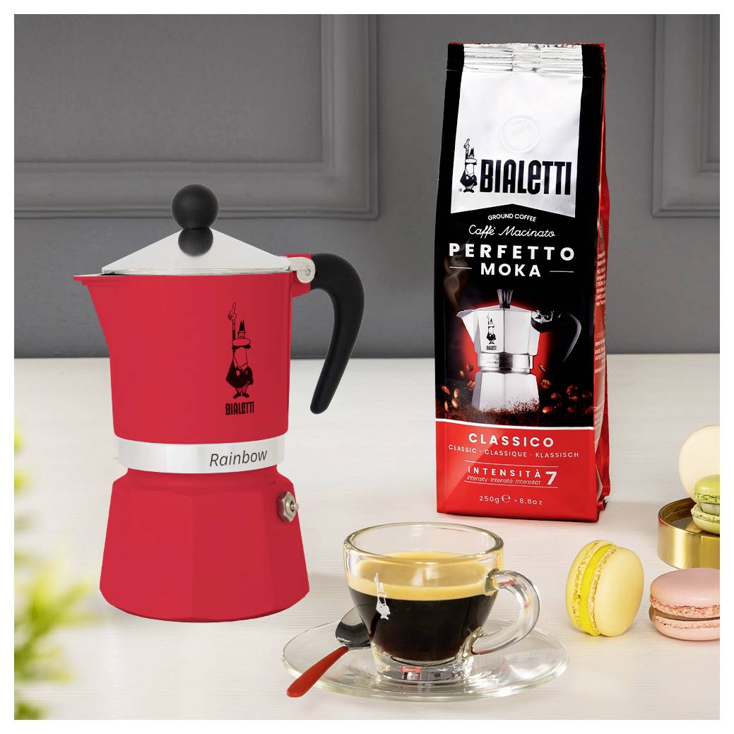 Ein roter Mokkakocher neben einer Tüte Bialetti Perfetto Moka Kaffee, einer Glasstasse Espresso und Macarons auf einer Küchenarbeitsplatte.