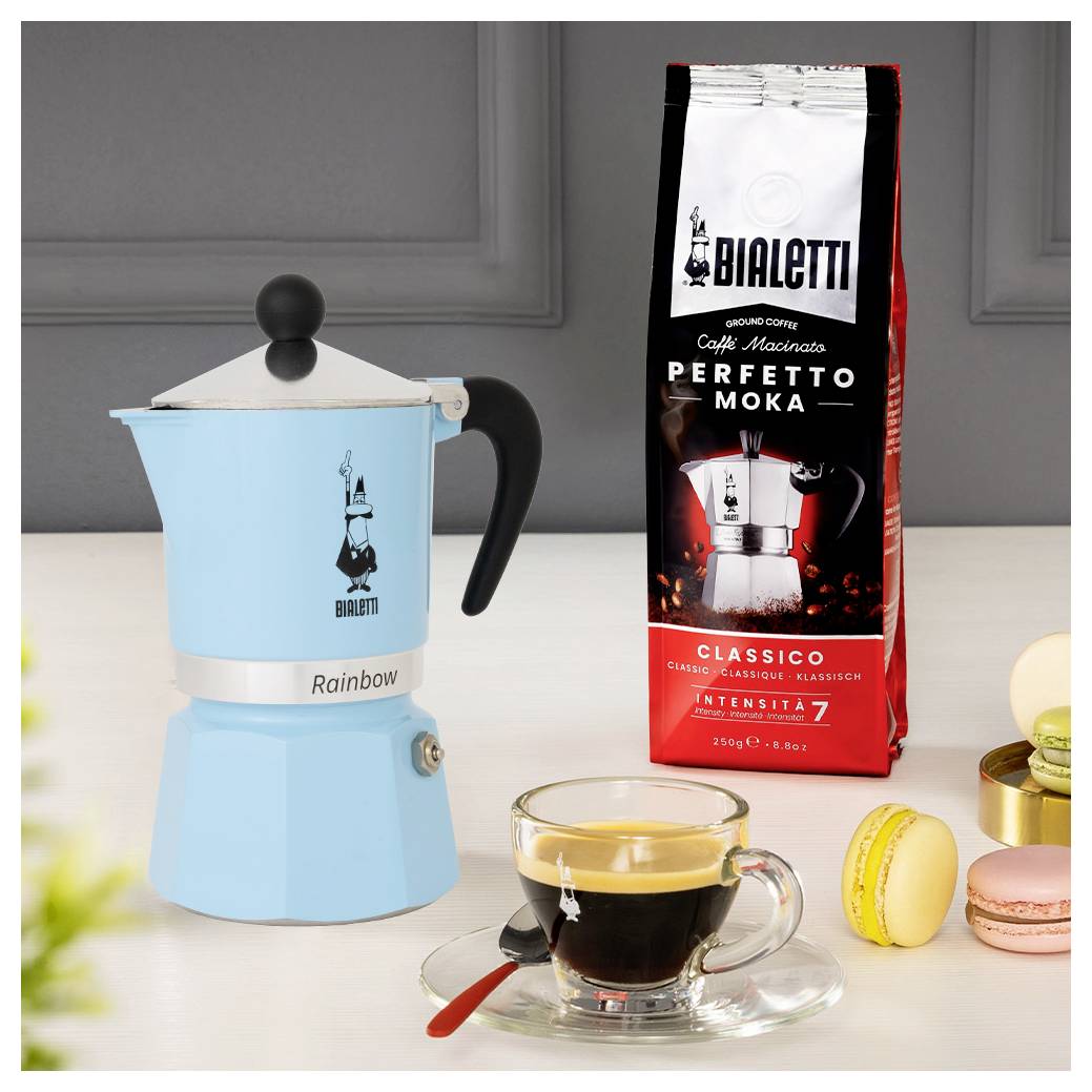 Ein blauer Mokkakocher, ein rotes Bialetti Perfetto Moka Kaffeepäckchen und ein Espresso in einer Glasstasse stehen auf einer Arbeitsplatte vor einer grauen Wand.