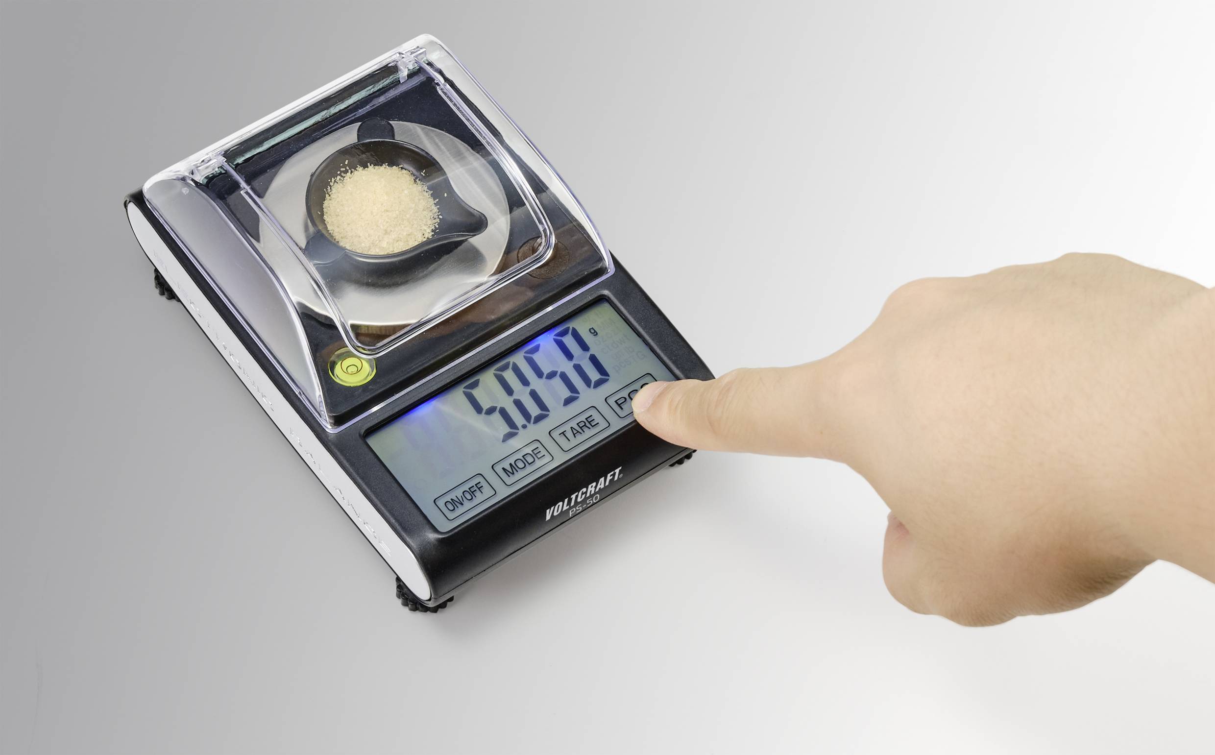 Eine digitale Waage zeigt ein Gewicht von 5,859 Gramm an. Eine Hand tippt auf die 'TARE'-Taste. Auf der Waage liegt ein kleiner Behälter mit Pulver.