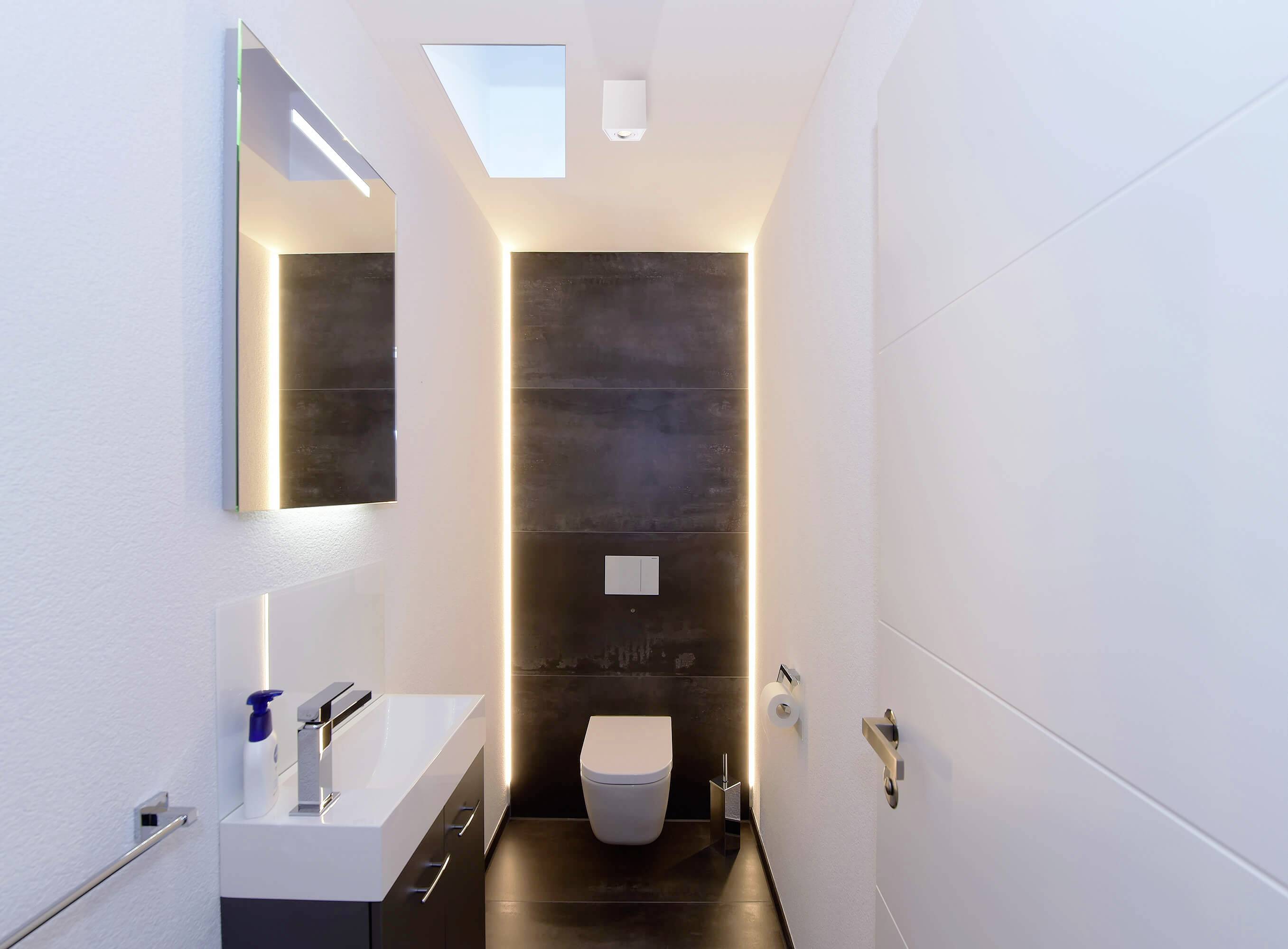 Modernes Badezimmer mit wandmontierter Toilette, Waschbecken und Spiegel. Dezente Beleuchtung betont den schwarzen Fliesenhintergrund.