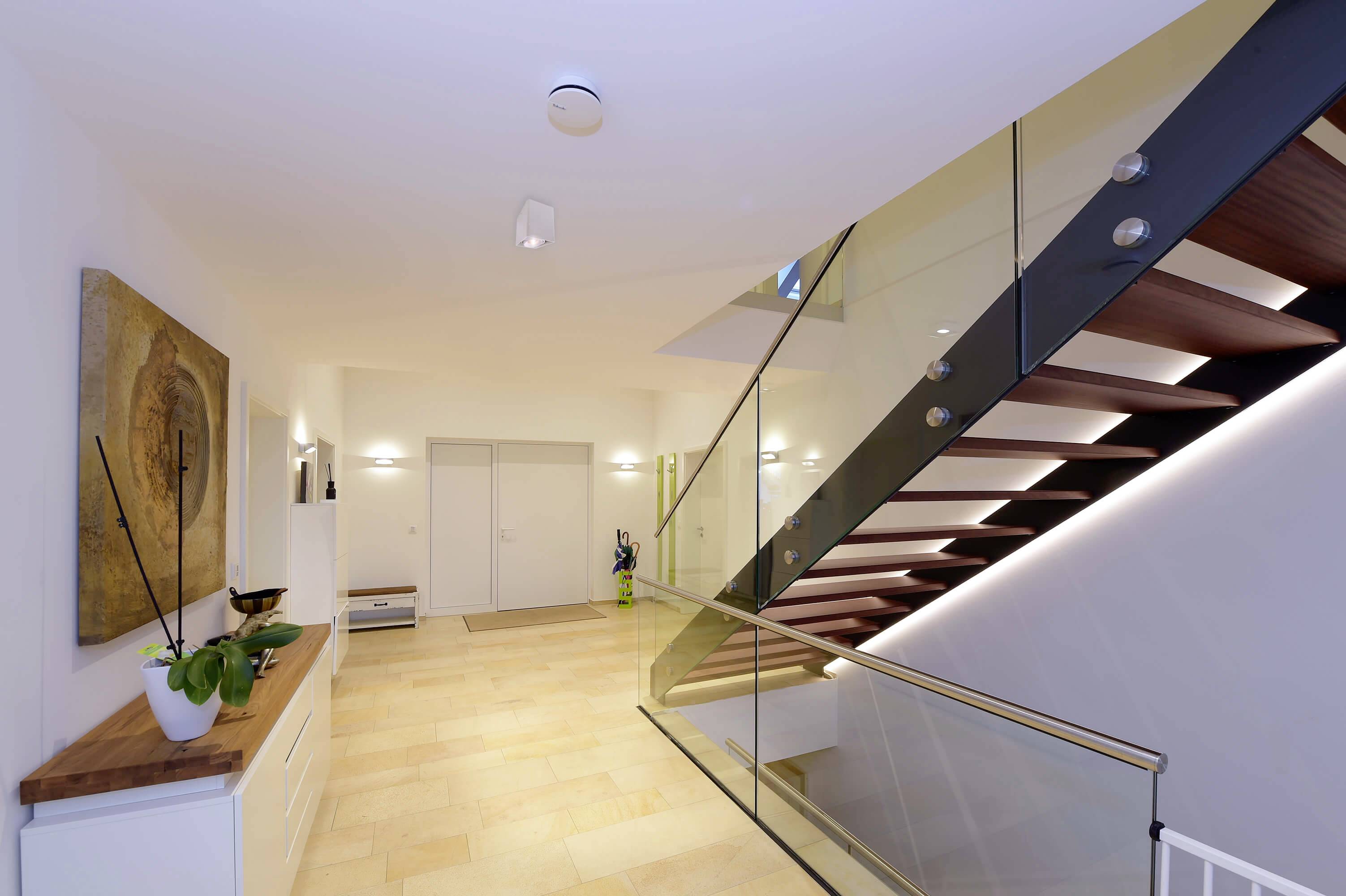 Wohnbereich mit cremefarbenem Fliesenboden, moderner Treppe aus dunklem Holz und Glasgeländer. An der Wand hängt ein abstraktes Kunstwerk.