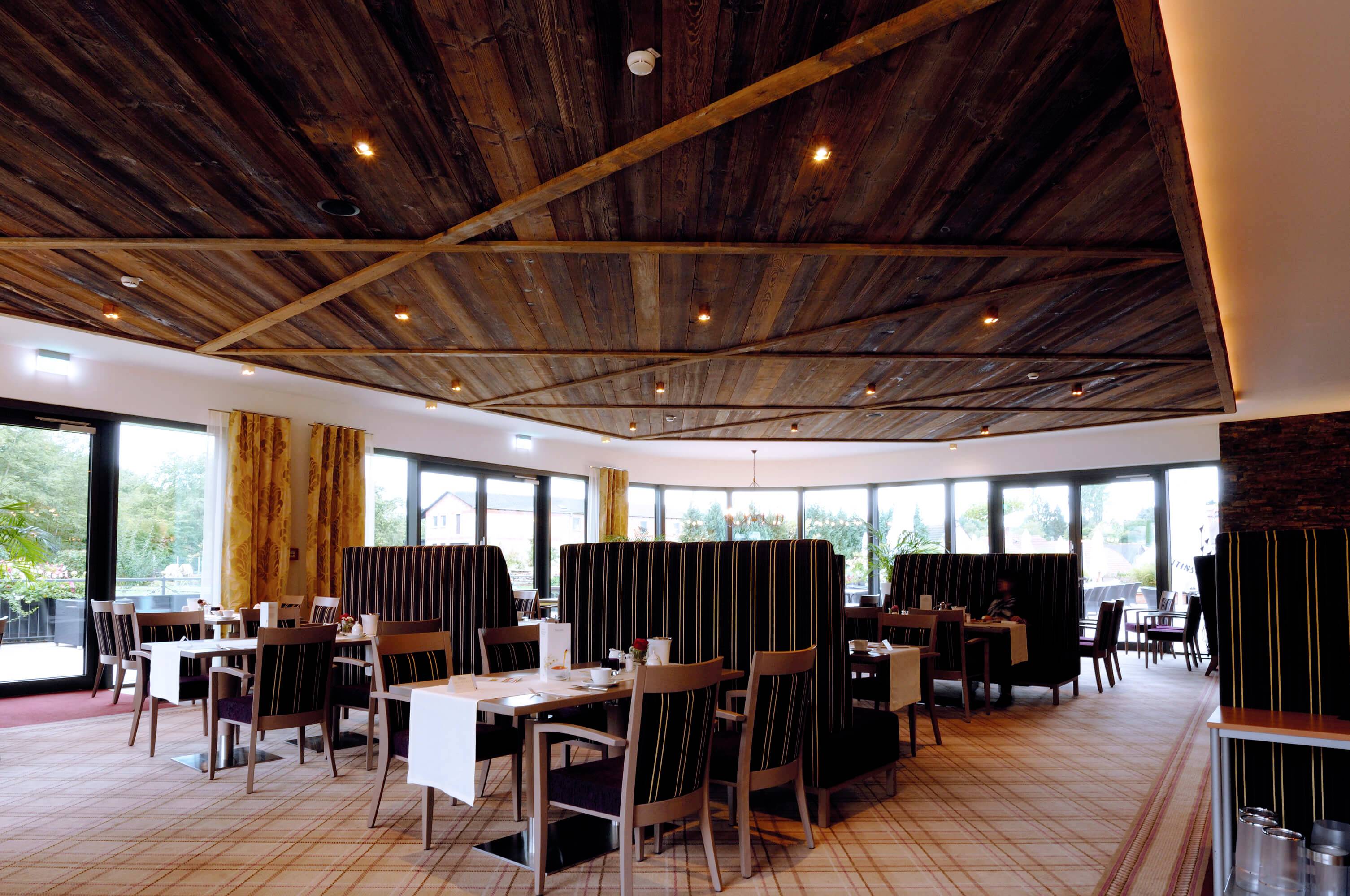 Ein modernes Restaurant mit Holzelementen an Decke und Wänden, leeren Tischen und gepolsterten Sitzbereichen, großen Fenstern mit Blick ins Freie.