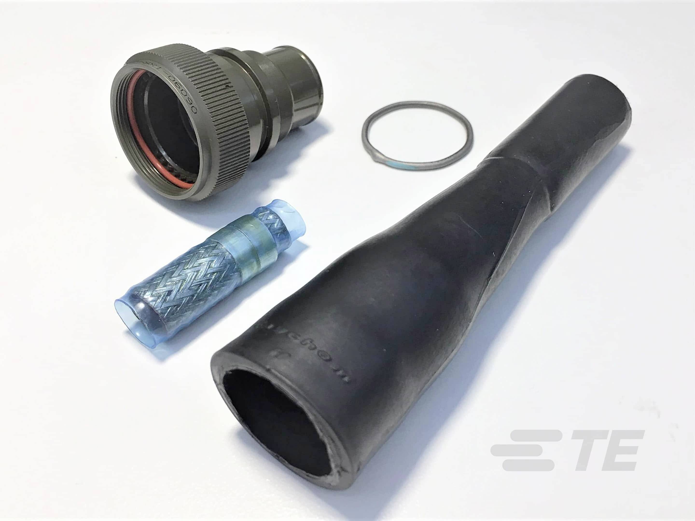 Ein militärischer Steckverbinder, ein Dichtungsgummi, eine bläuliche Hülle und ein Kabelschutz liegen auf einer weißen Fläche.