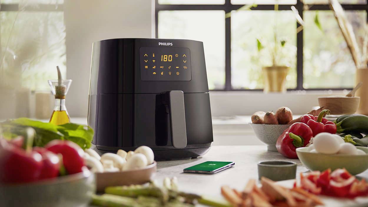 Ein schwarzer Heißluftfritteuse mit digitalem Display zeigt '180°'. Umgeben von frischem Gemüse und einem Smartphone auf der Küchenarbeitsplatte.