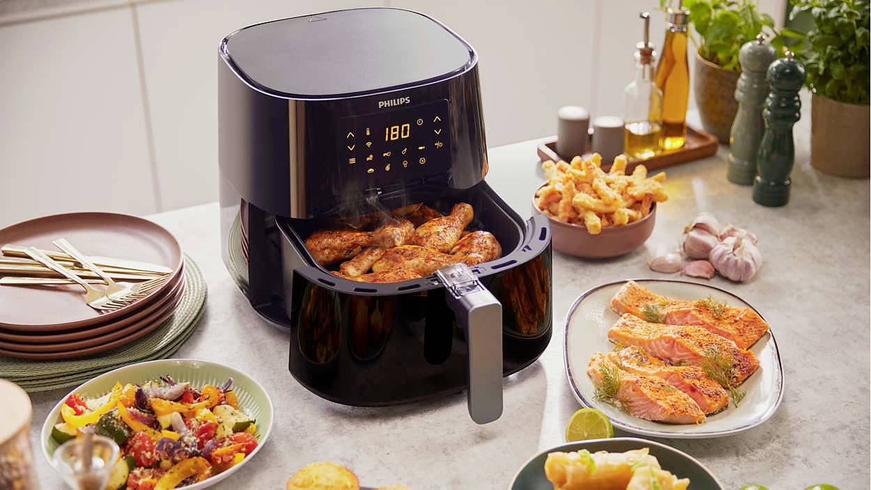 Ein Airfryer auf einem Tisch bereitet Hähnchenflügel zu. Umgeben von verschiedenen zubereiteten Speisen wie Lachs und Pommes.