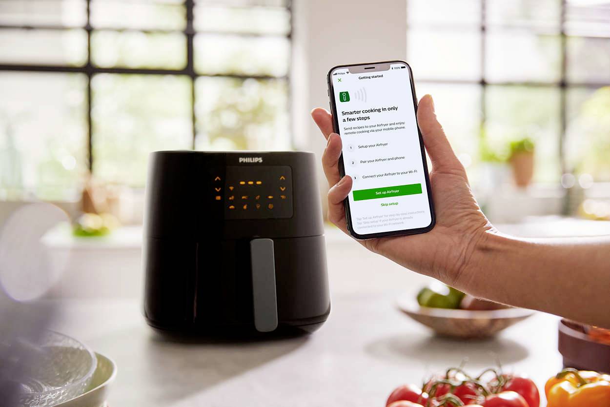 Eine Person hält ein Smartphone mit einer App, die mit einer schwarzen Philips-Heißluftfritteuse in einer modernen Küche verbunden ist.
