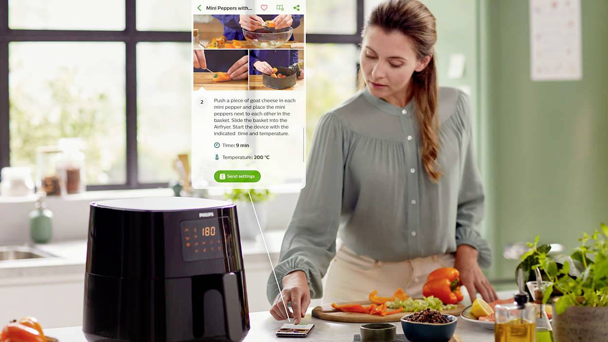 Frau in der Küche kocht mit einer Heißluftfritteuse. Auf dem Handy ist eine Rezeptanleitung zu sehen. Gemüse liegt auf dem Tisch.