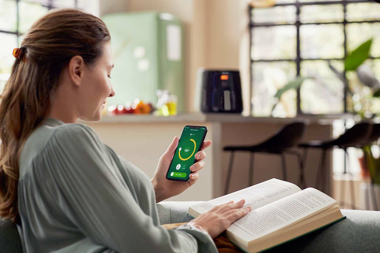 Eine Frau sitzt auf einem Sofa, hält ein Smartphone mit einer grünen Gesundheits-App-Anzeige und liest ein Buch; Küchenhintergrund.