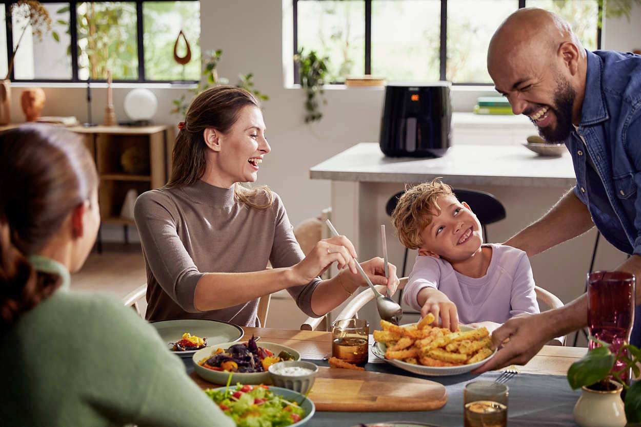 Eine Familie sitzt zusammen am Tisch und genießt eine Mahlzeit mit Pommes und Salat. Ein Mann serviert lächelnd das Essen.