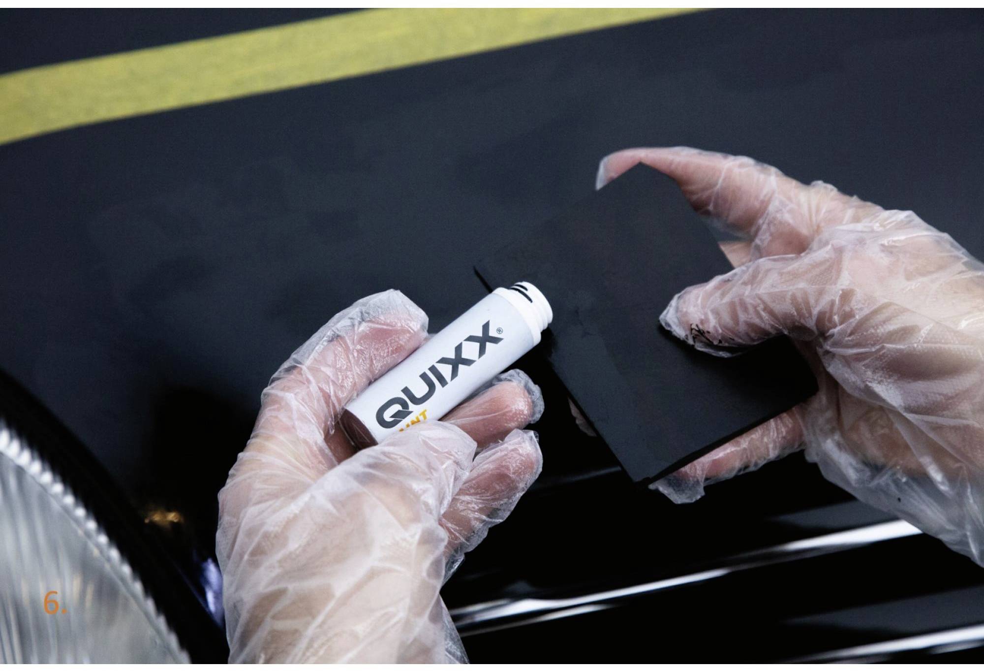 Eine Person trägt Quixx-Politur mit einem Tuch auf eine Autokarosserie auf. Die Hände sind in Handschuhen geschützt.