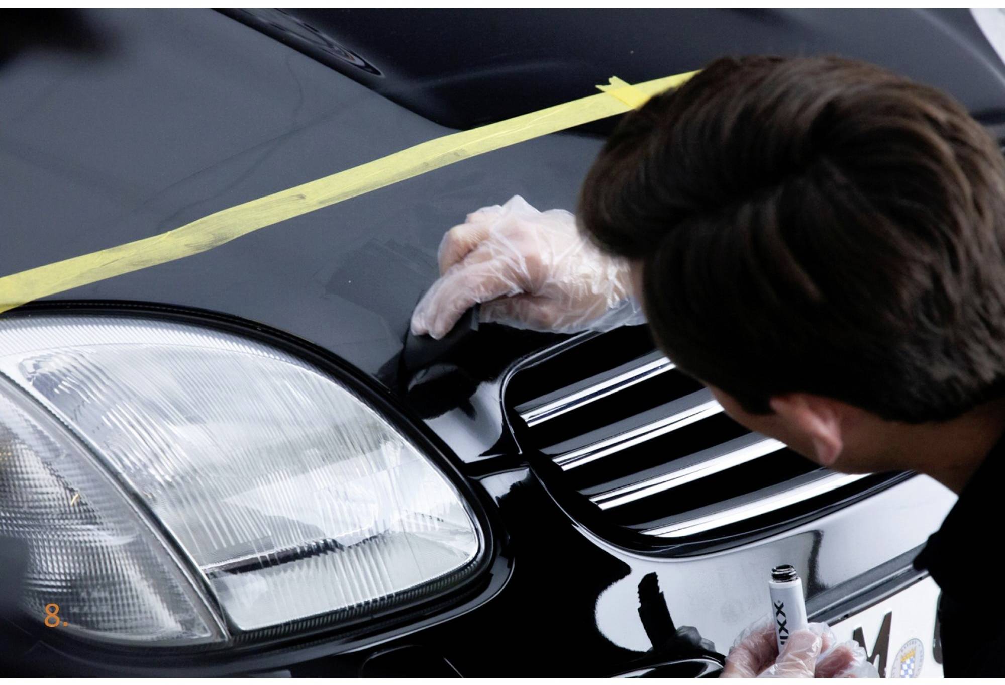 Eine Person trägt mit Handschuhen vorsichtig Politur auf die Motorhaube eines schwarzen Autos auf, um Kratzer zu entfernen.