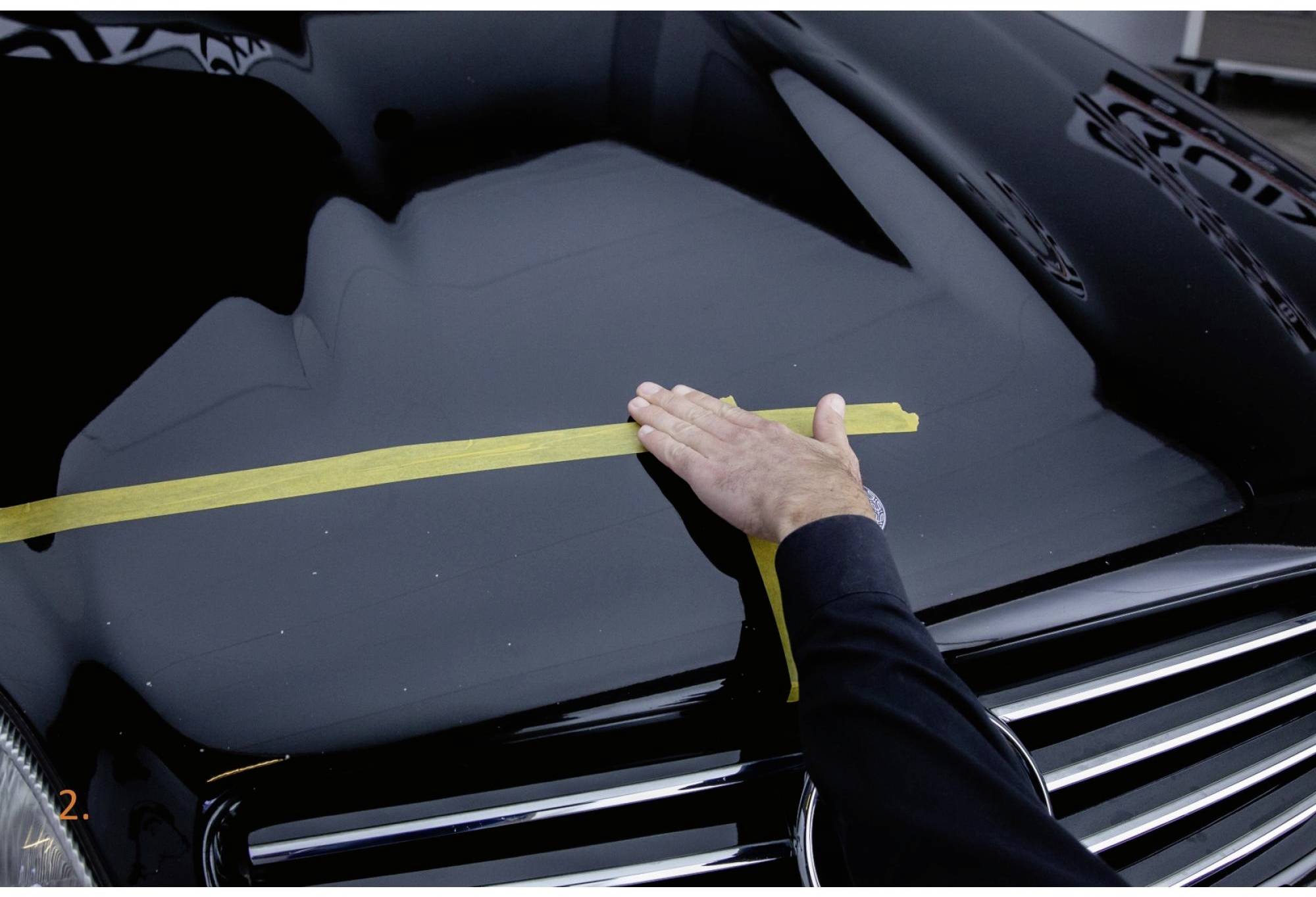 Ein Mensch legt gelbes Klebeband auf die Motorhaube eines schwarzen Autos, um eine gerade Linie zu markieren.
