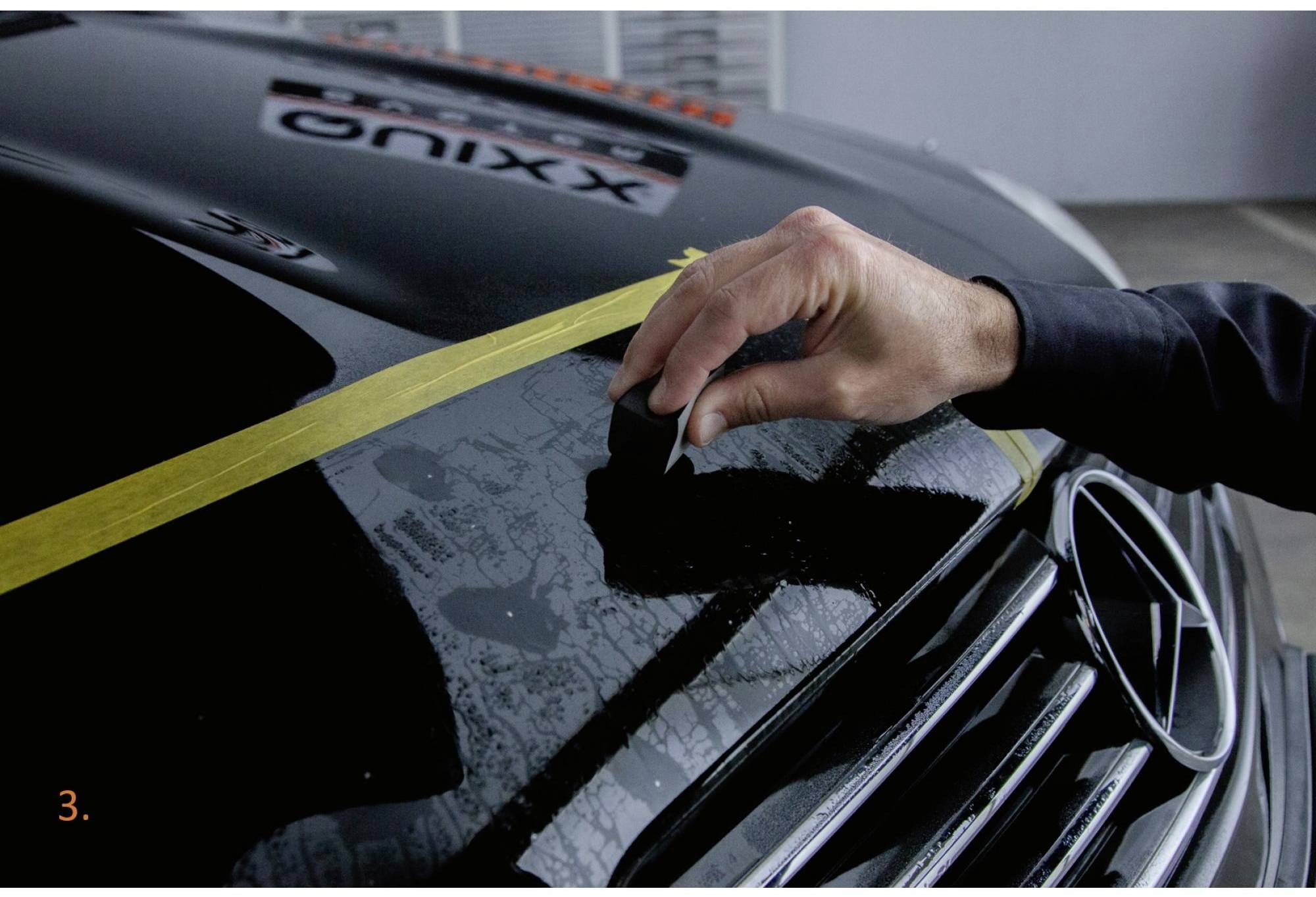 Hand poliert die Motorhaube eines schwarzen Autos mit gelbem Klebeband darauf. Mercedes-Logo sichtbar.