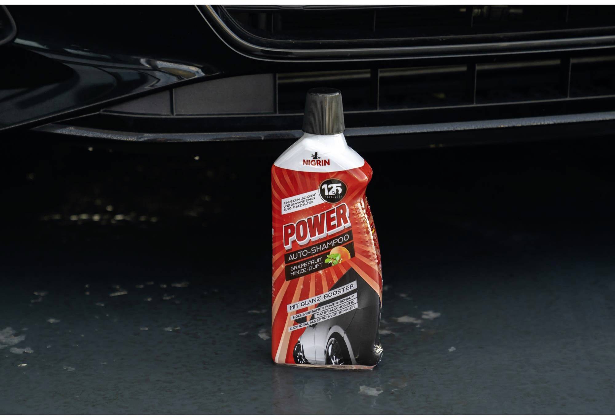 Eine Flasche roten Auto-Shampoos steht auf einem nassen Boden vor einem Autostoßfänger. Die Flasche hat ein schwarzes Verschluss.