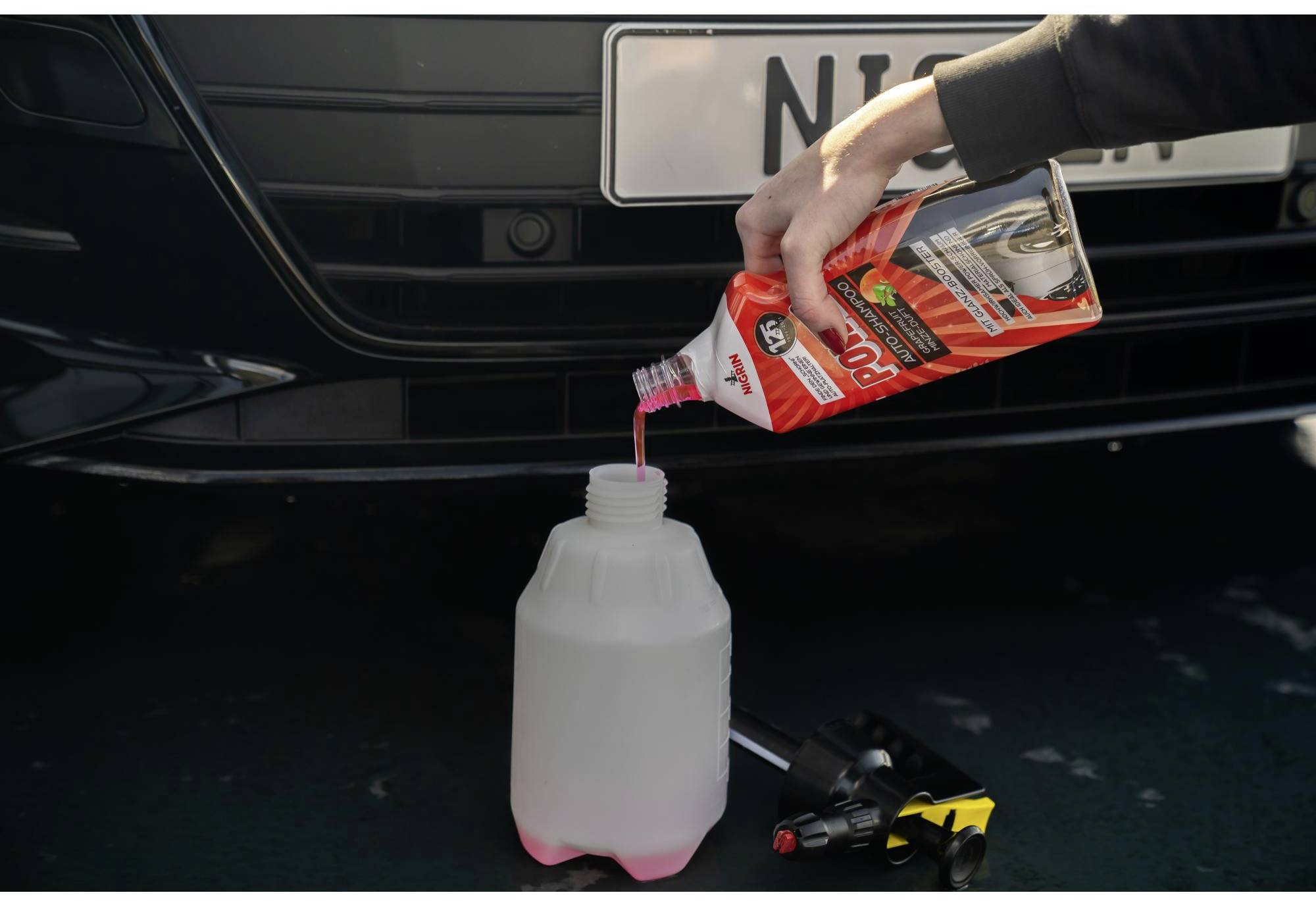 Eine Person gießt rosa Flüssigkeit aus einem Kanister in einen großen Kanister unter der Motorhaube eines schwarzen Autos.
