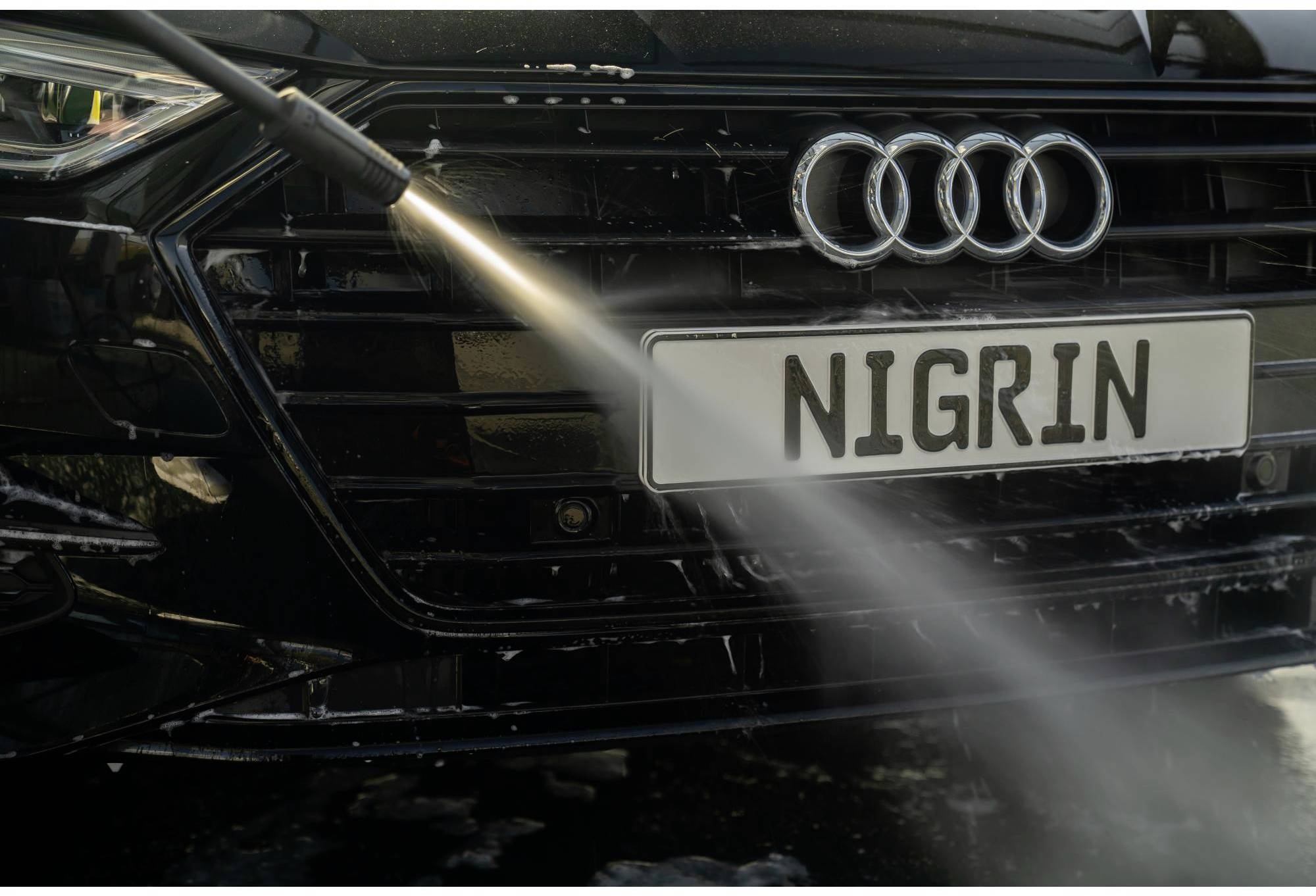 Ein schwarzes Auto wird mit einem Hochdruckreiniger gereinigt. Der Fokus liegt auf dem Kühlergrill und dem Scheinwerfer.