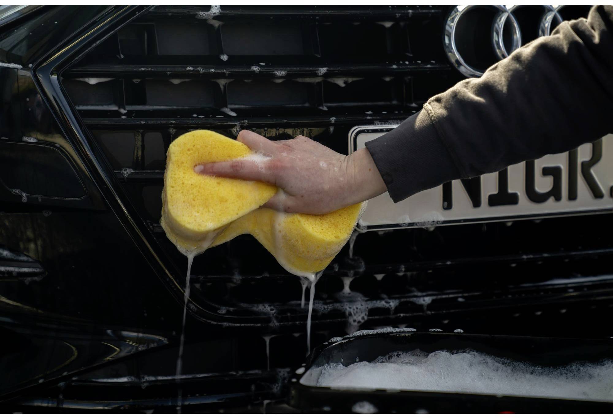 Eine Person wäscht den Kühlergrill eines Autos mit einem gelben Schwamm. Schaum und Wasser sind sichtbar, Nummernschild teilweise verdeckt.