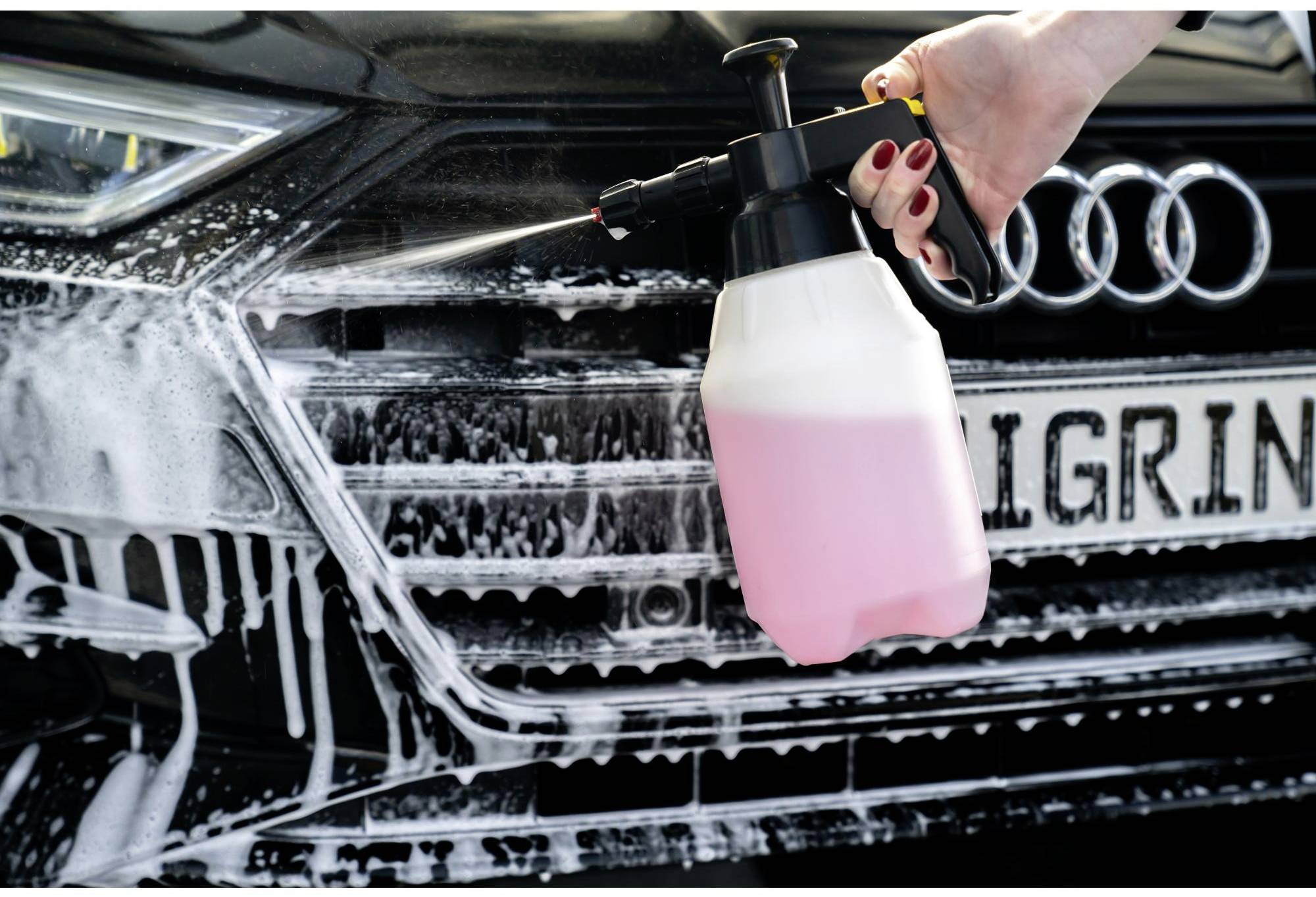 Eine Hand sprüht mit einem Reinigungsgerät eine pinke Flüssigkeit auf die Front eines Autos, das mit Schaum bedeckt ist.