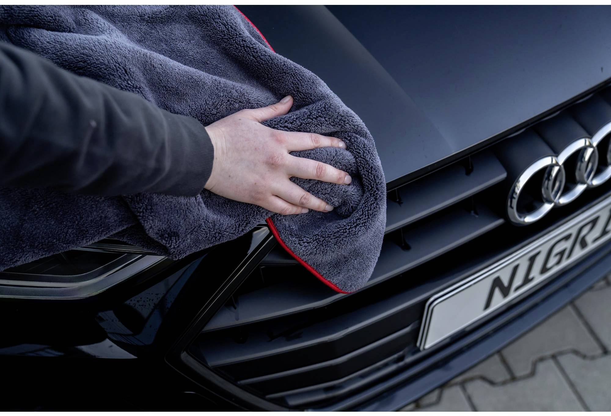 Eine Person reinigt mit einem grauen Tuch den Kühlergrill eines schwarzen Autos.