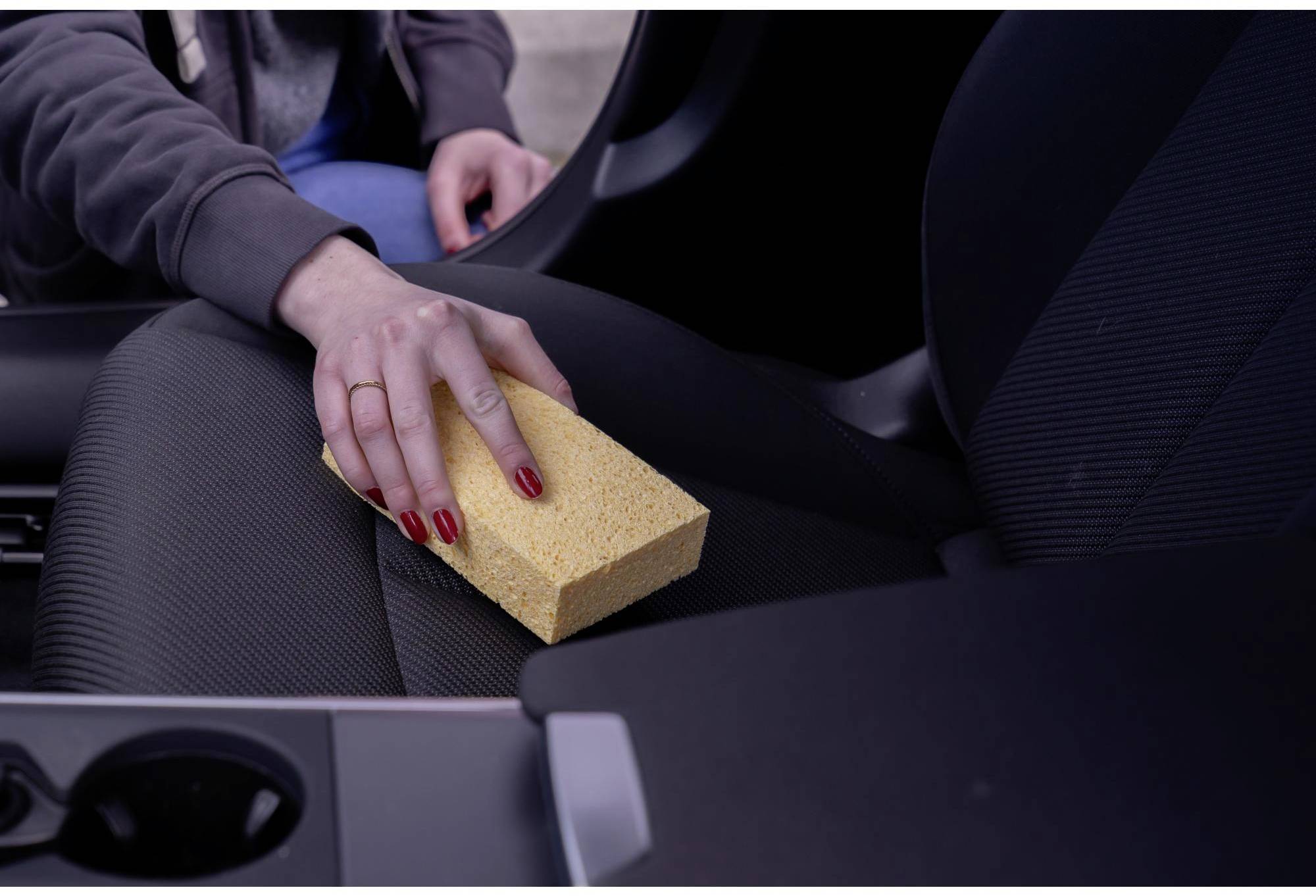 Eine Person reinigt den Autositz mit einem gelben Schwamm. Fokus liegt auf dem Schwamm und der Hand.