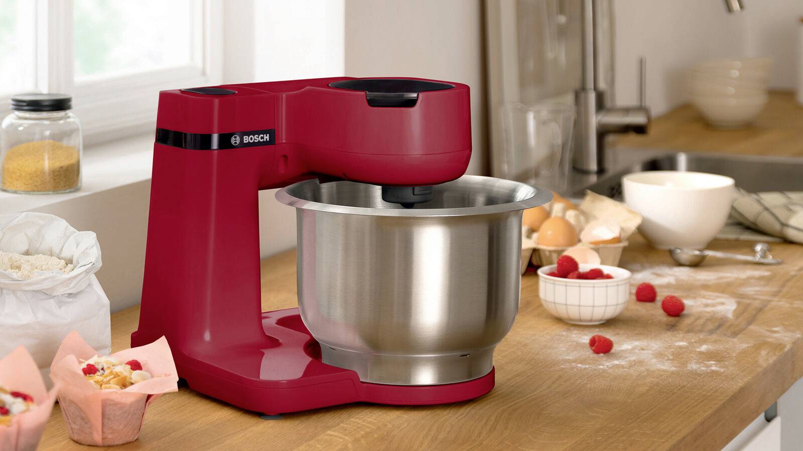 Rote Küchenmaschine mit Edelstahl-Rührschüssel auf Holzarbeitsplatte. Neben der Maschine liegen Mehl, Eier und Himbeeren.