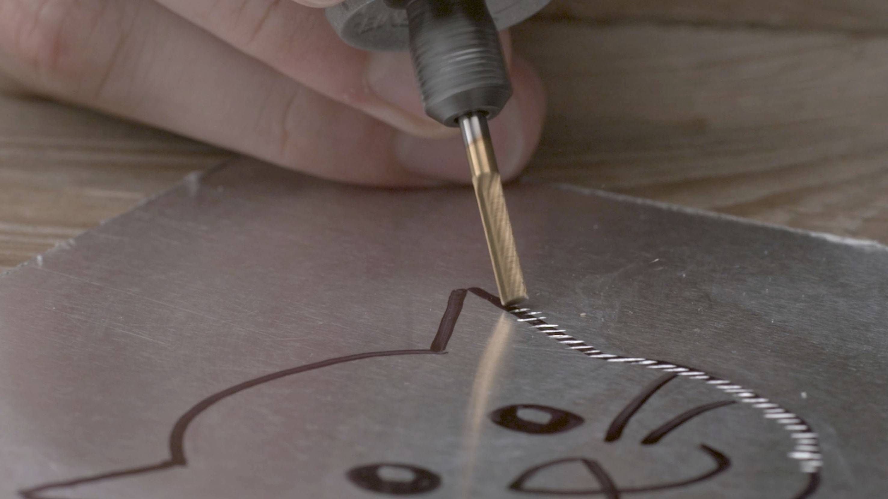 Eine Nahaufnahme zeigt, wie eine Hand mit einem Metallwerkzeug die Umrisse einer Katze auf eine Metallplatte graviert.