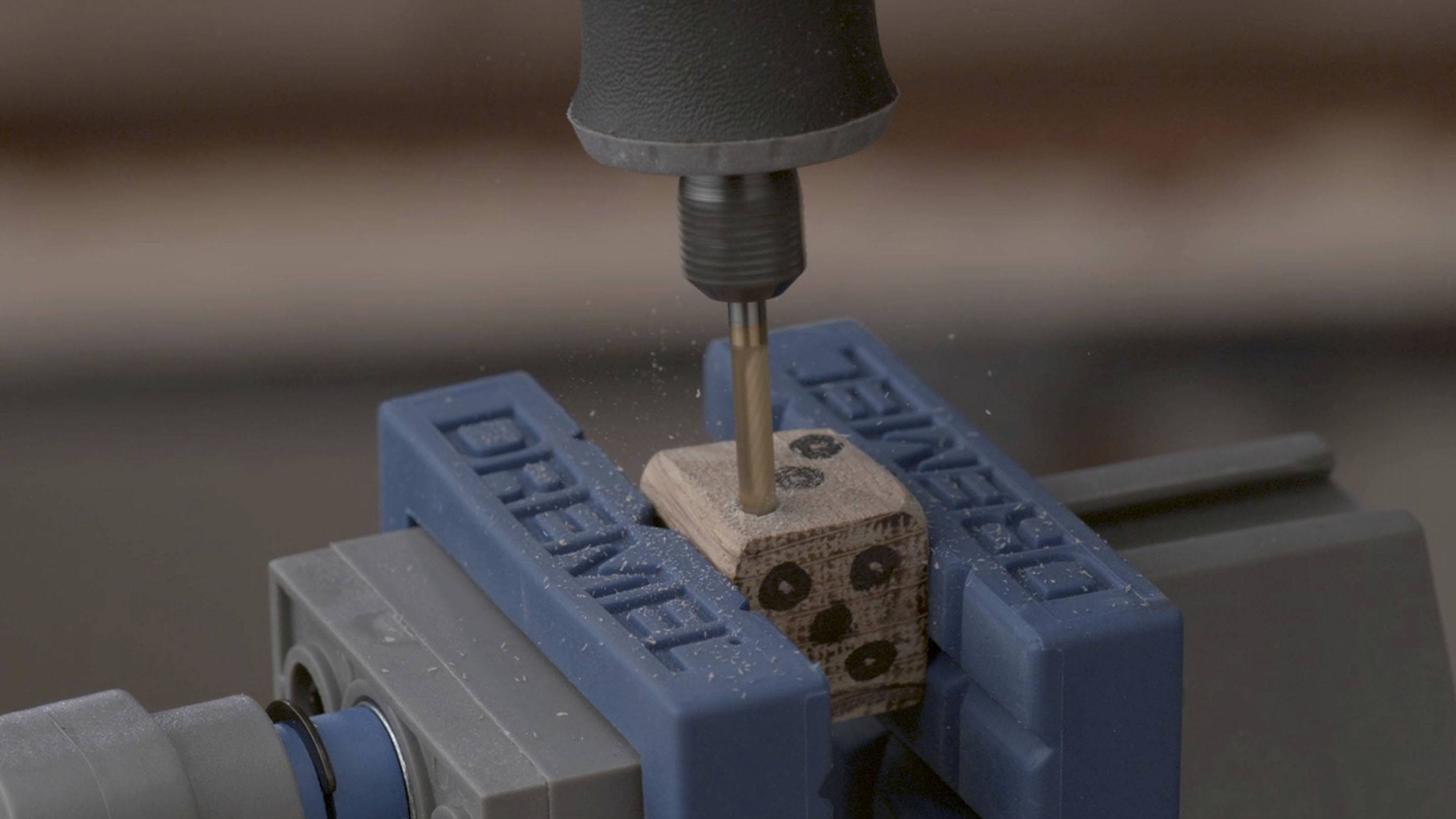 Eine Fräsmaschine bearbeitet einen kleinen Holzblock, der in einem Schraubstock eingespannt ist. Holzspäne fliegen auf.