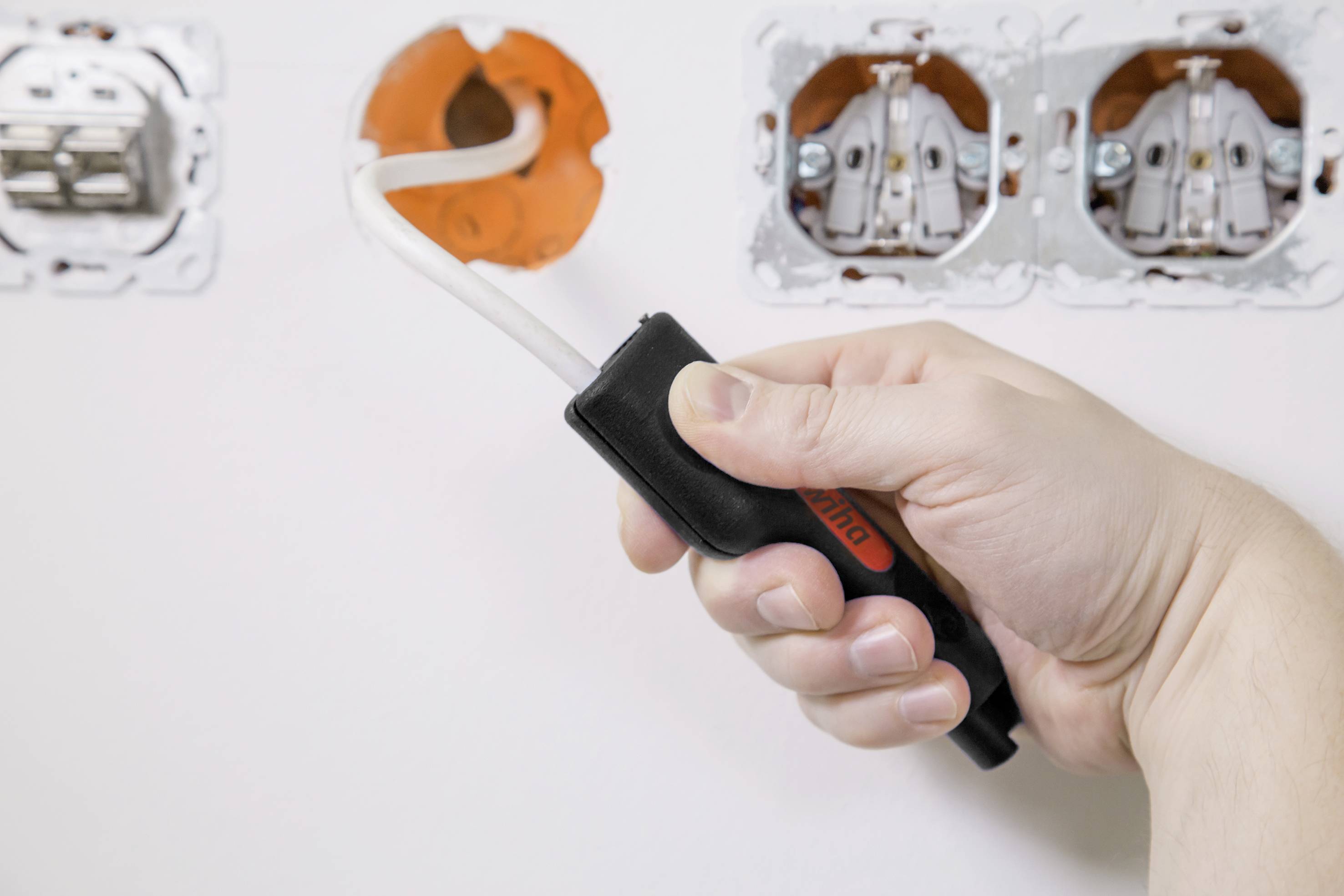 Eine Hand hält eine Spannungsprüfer in der Nähe von offenen Steckdosen in einer weißen Wand, während elektrische Arbeiten durchgeführt werden.