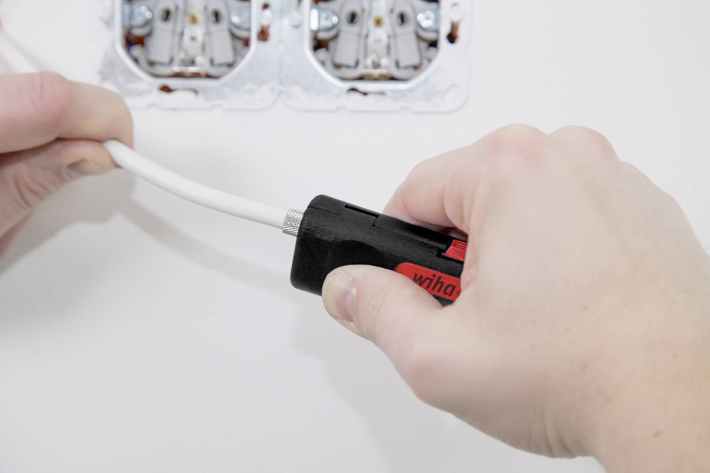 Eine Hand hält ein Werkzeug, das einen Kabelmantel von einem Elektrokabel abisoliert. Im Hintergrund sind Steckdosen erkennbar.